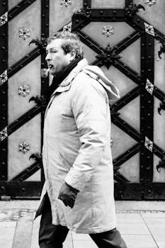 Man in coat walks past an ornate metal door in a city street, winter.