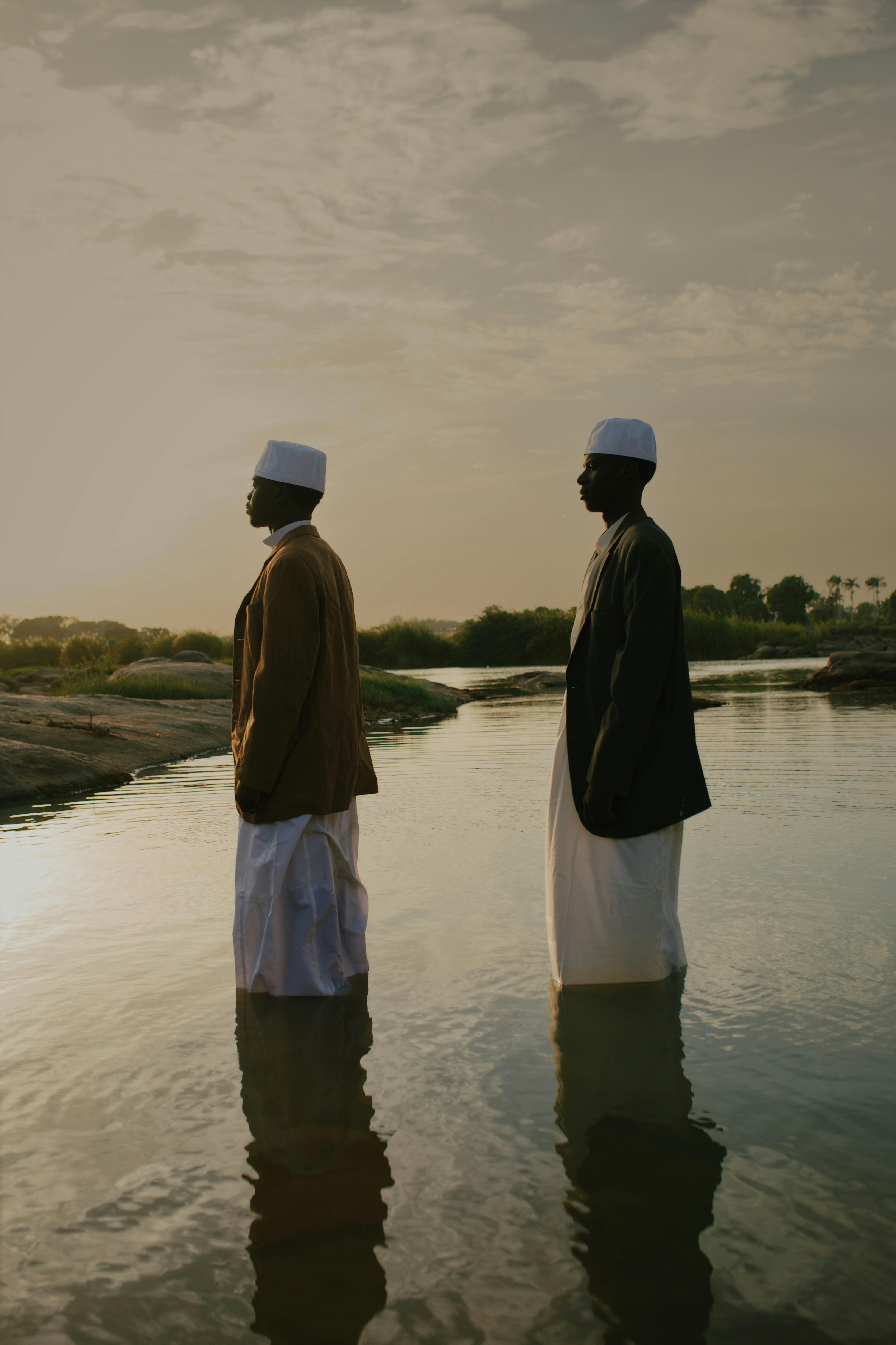 Gratis Due uomini in abiti tradizionali sono in piedi in un fiume placido al tramonto, che riflette il tranquillo paesaggio della Nigeria. Foto a disposizione