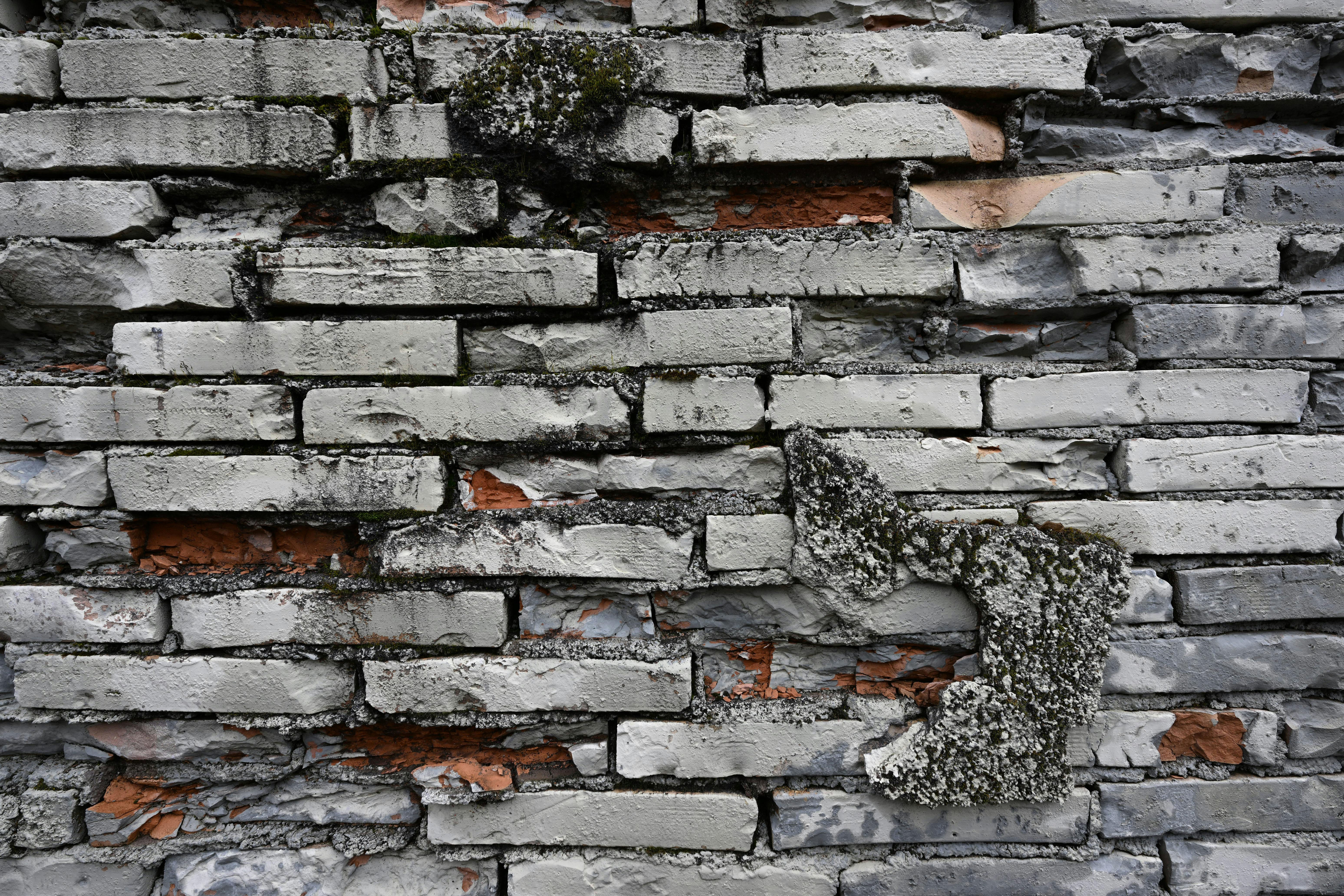 Deteriorated brick wall with crumbling mortar joints, cracked and spalling bricks, and moss growth from long-term moisture infiltration