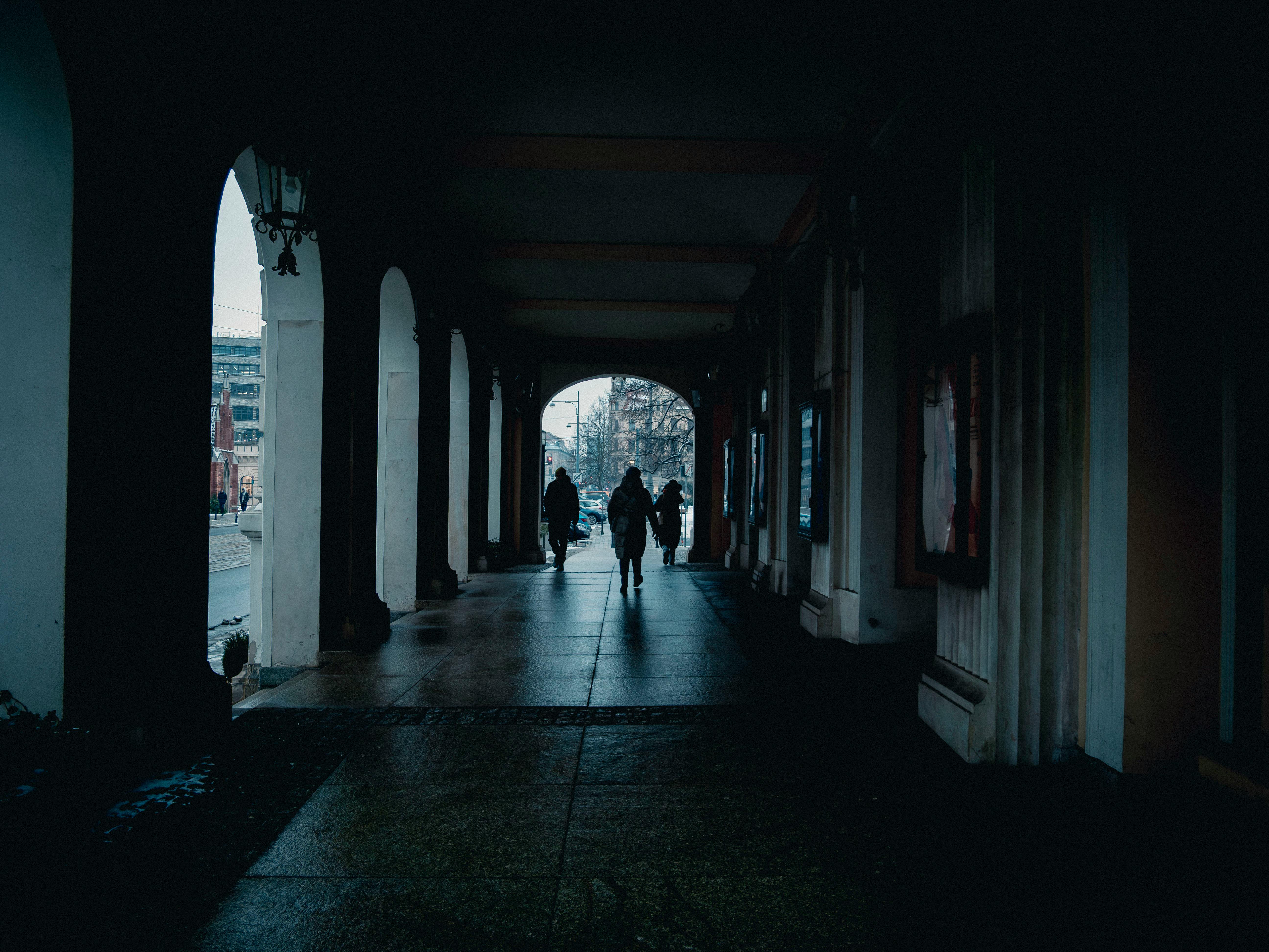 Gratis Orang-orang berjalan di bawah lengkungan gelap, menciptakan siluet dramatis di latar belakang kota. Foto Stok