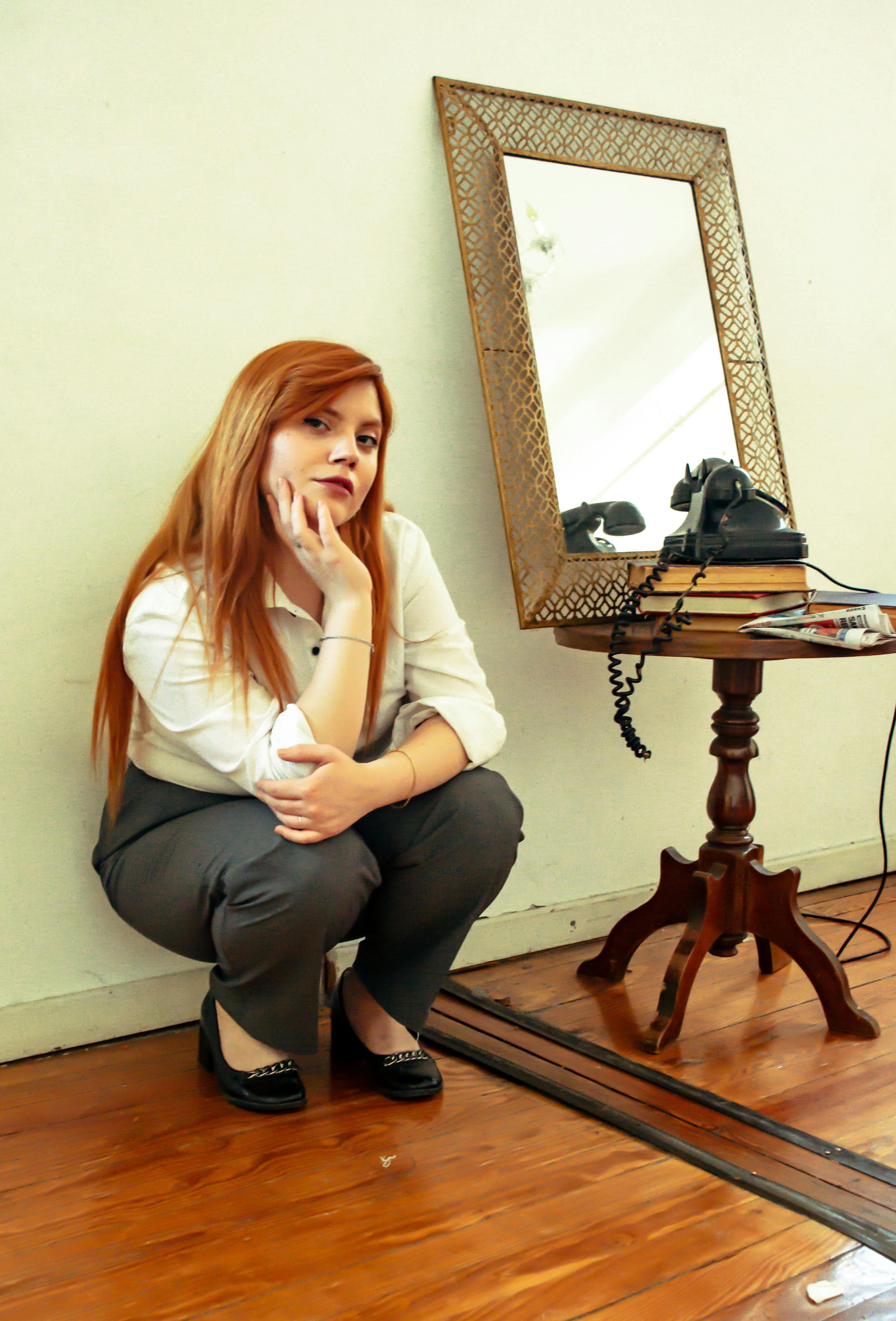 Bezpłatne Młoda kobieta kuca przy zabytkowym lustrze i telefonie w pomieszczeniu. Zdjęcie z galerii