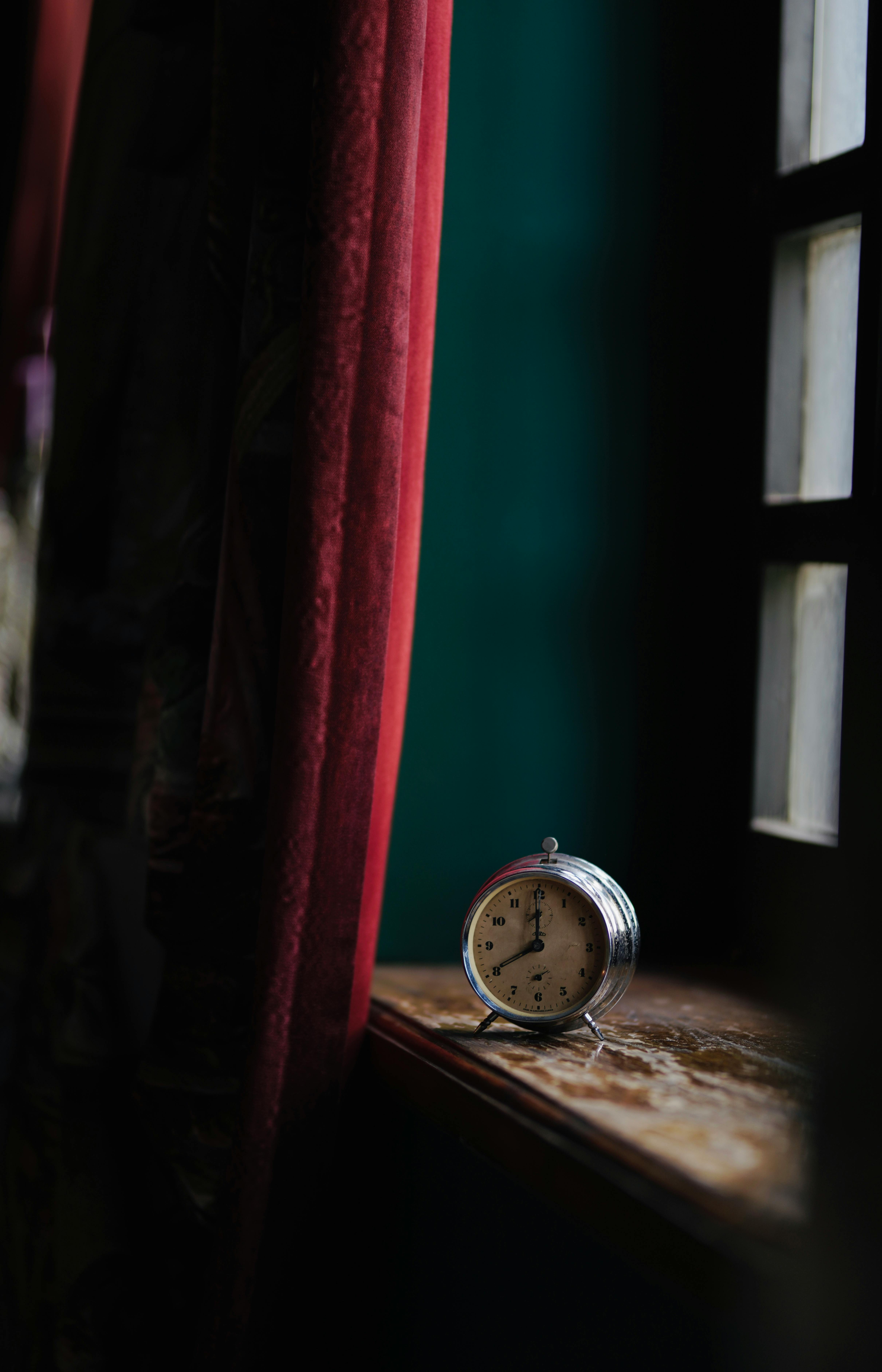 Gratis Una sveglia d'epoca è appoggiata sul davanzale di una finestra, incorniciata da tende rosse, in una stanza scarsamente illuminata. Foto a disposizione