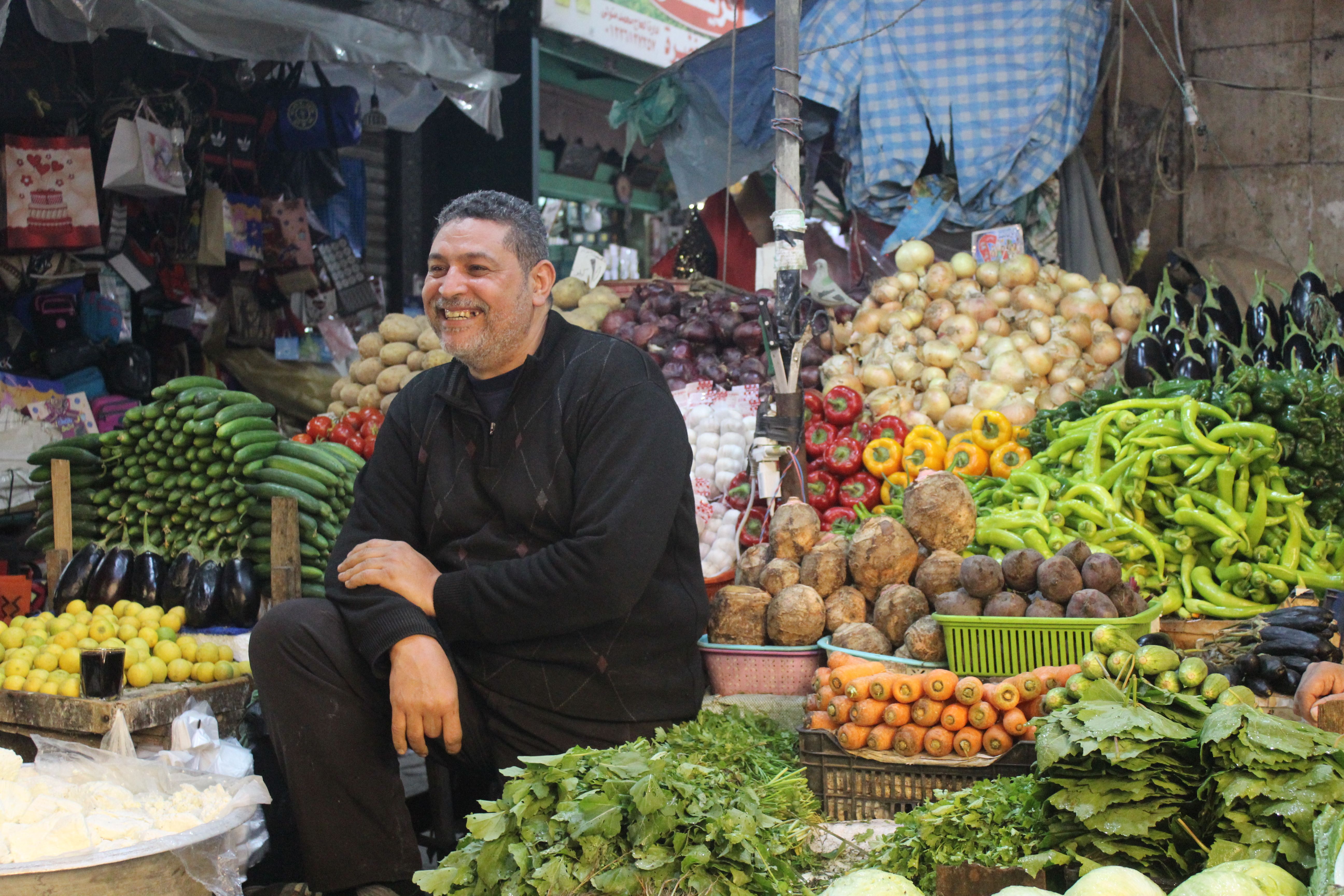 Smiling vendor amidst a colorful array of fresh produce at a bustling market.