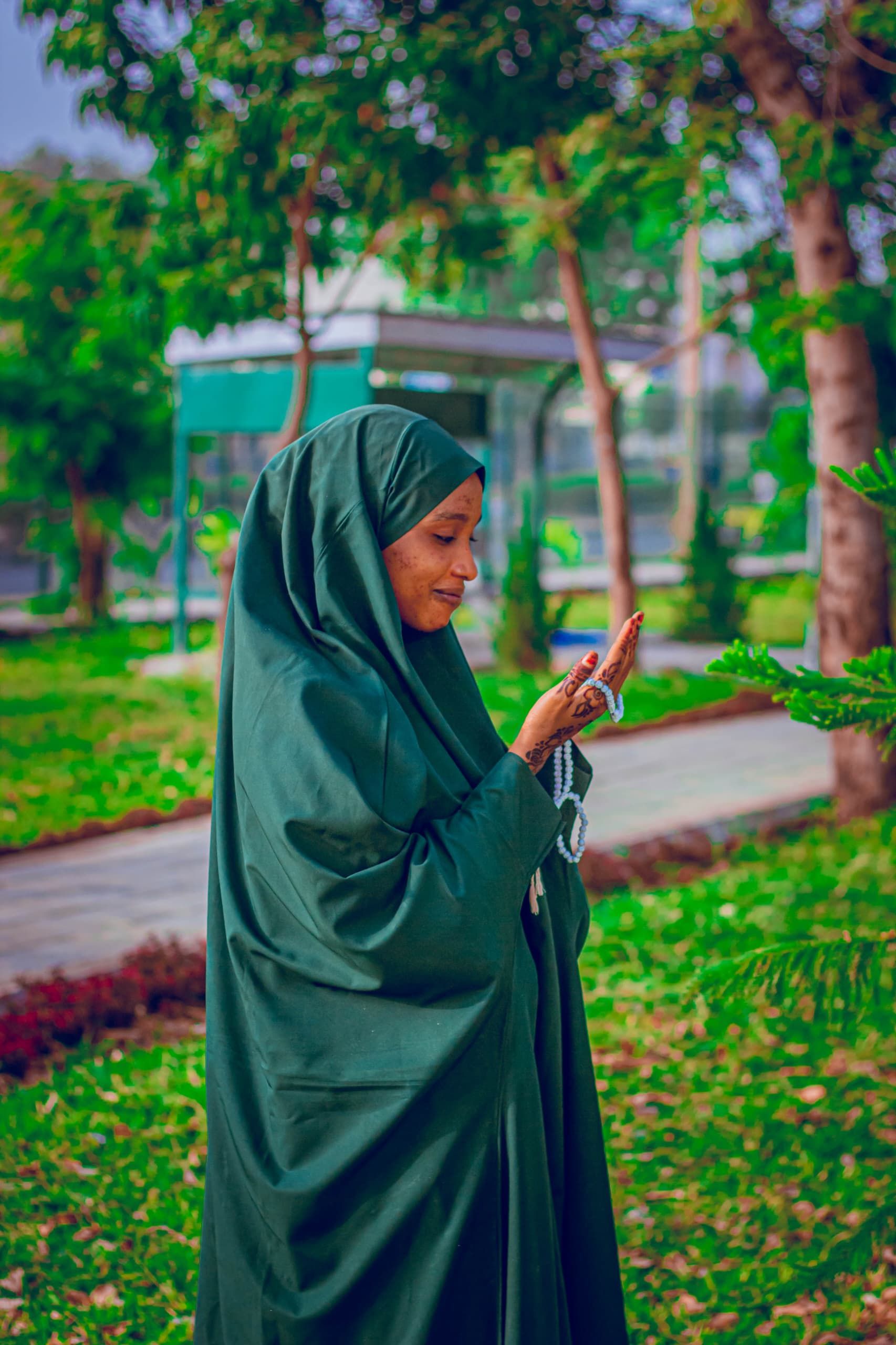 Woman in a green hijab standing outdoors, praying in a peaceful park setting.