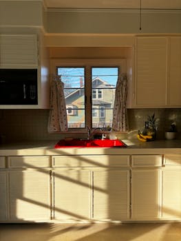 A cozy kitchen with sunlight streaming in, highlighting a red sink and floral curtains.