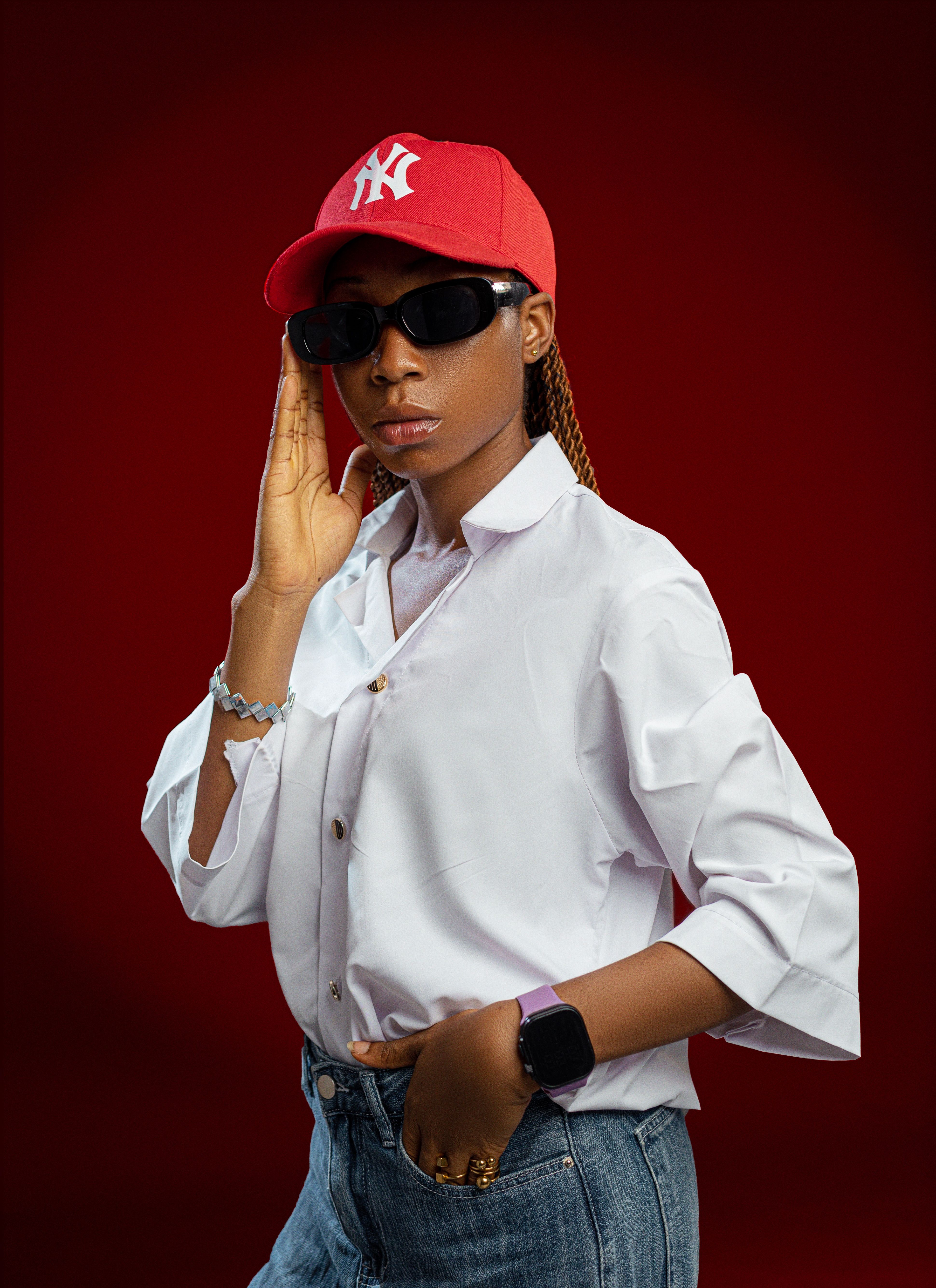 Confident woman posing with a red cap and sunglasses, striking a bold pose against a red backdrop.
