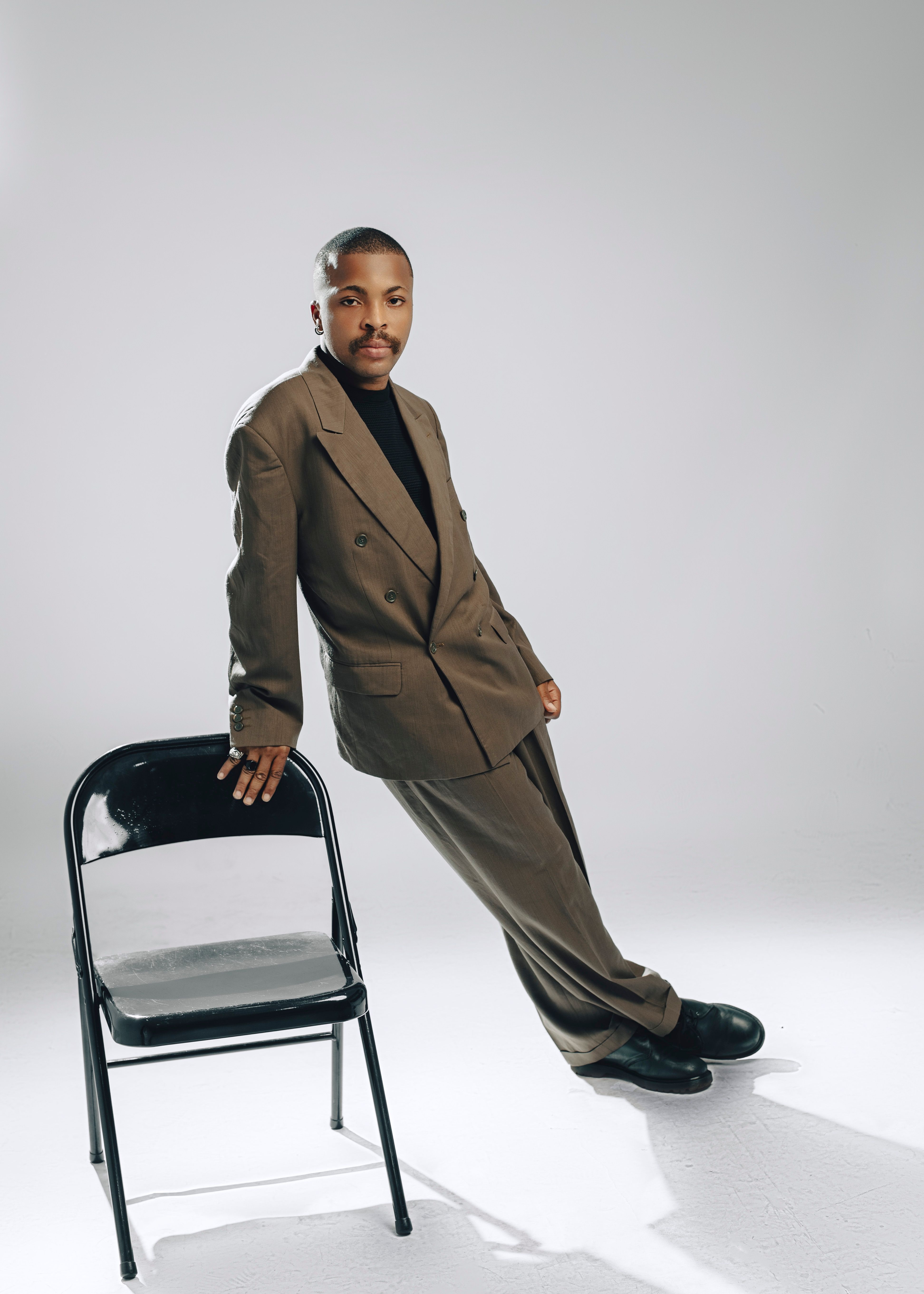 Elegantly dressed man leans on a chair in a studio setting, exuding confidence.