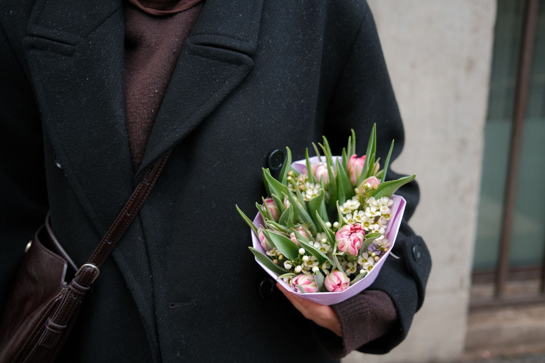 A person in a dark coat holds a bouquet of pink tulips and white flowers outdoors.