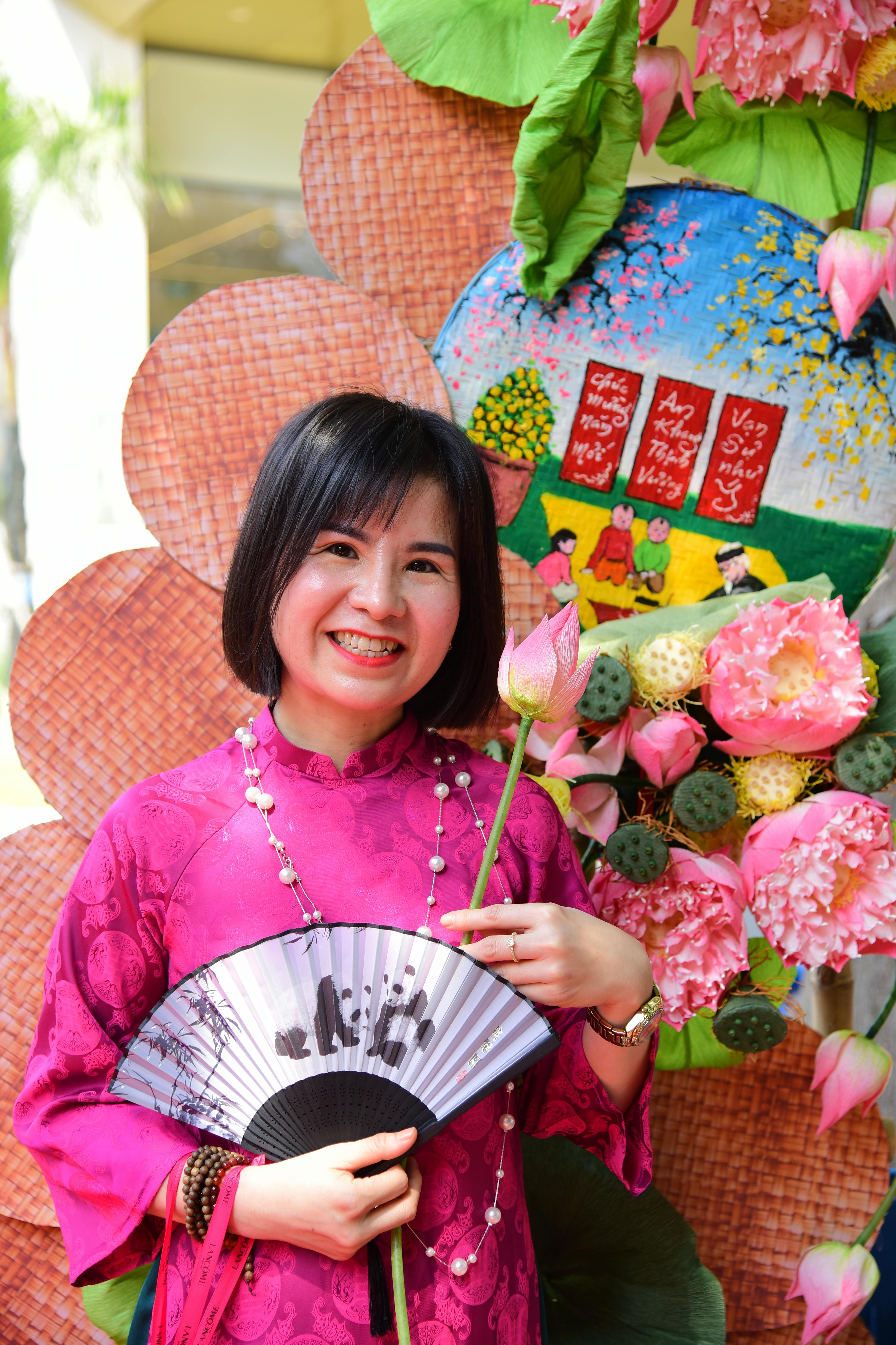 Gratis Donna sorridente in abito tradizionale che tiene in mano fiori di loto e un ventaglio, sullo sfondo decorazioni vivaci. Foto a disposizione