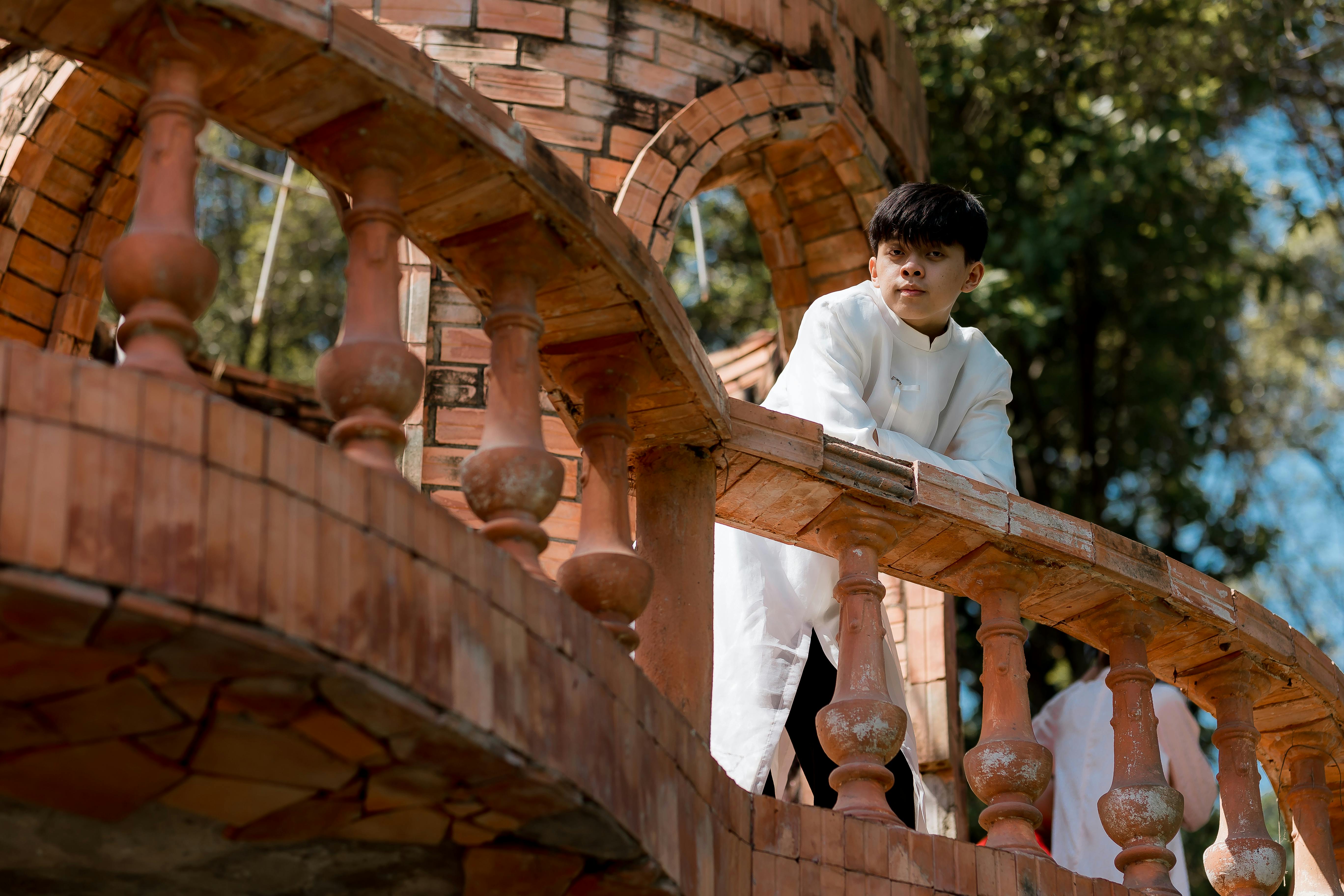 Ücretsiz Geleneksel kıyafetler giymiş genç bir adam, dışarıdaki rustik tuğla bir balkona yaslanmış duruyor. Stok Fotoğraflar