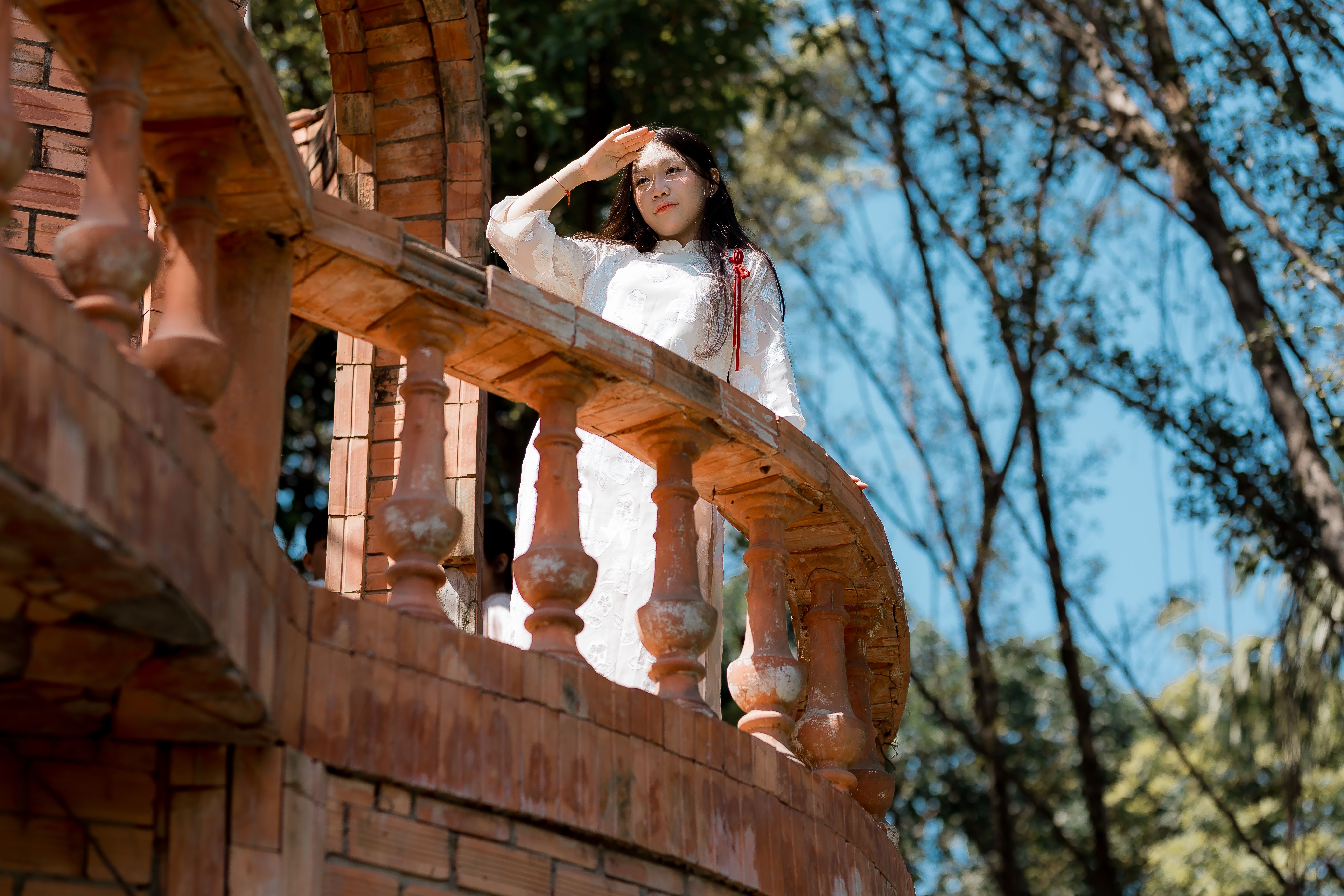 Woman in white dress enjoys a sunny day on a rustic balcony surrounded by nature.