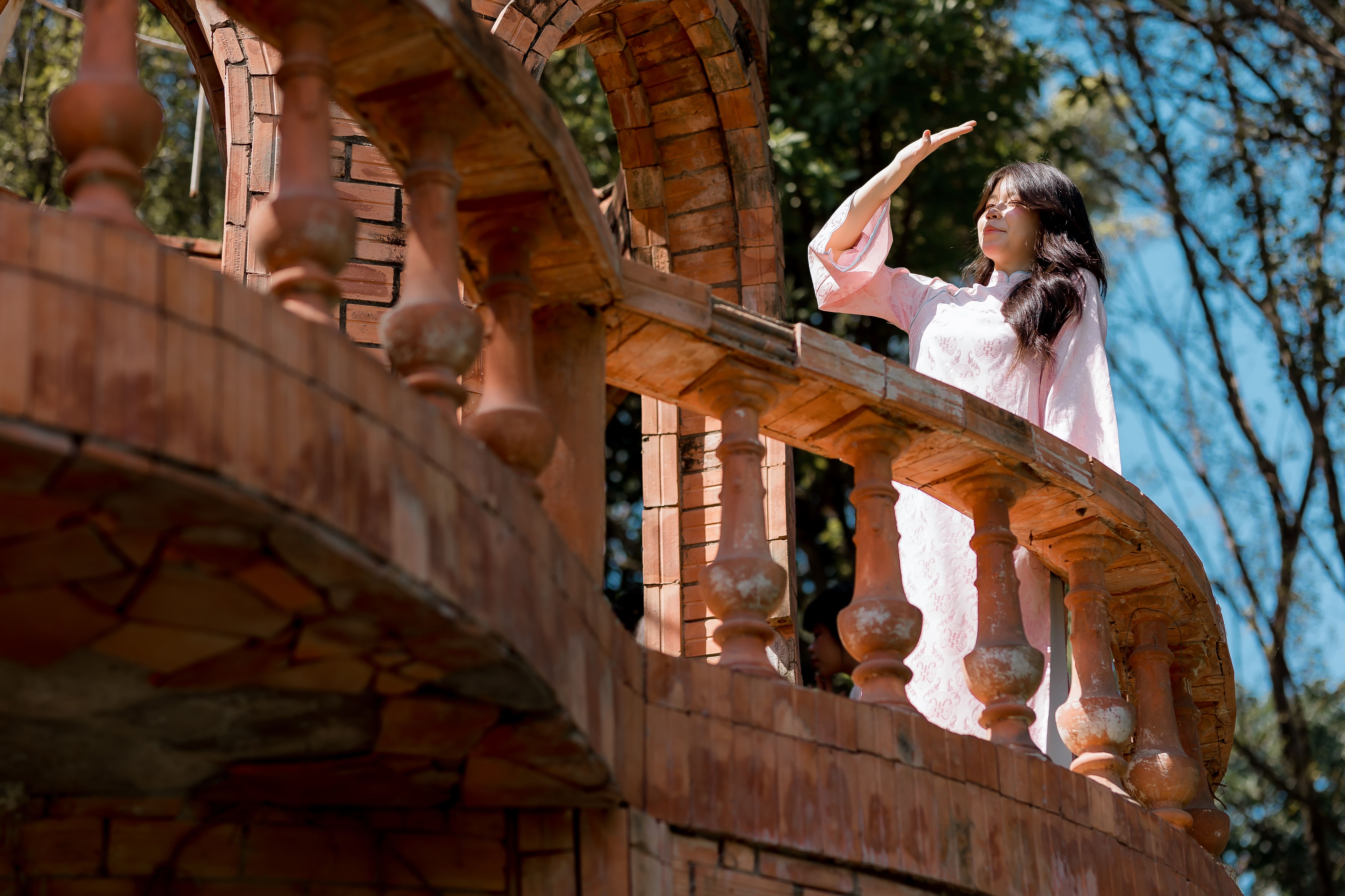 Young woman in a pink dress stands on a brick balcony under the sun, shielding her eyes.