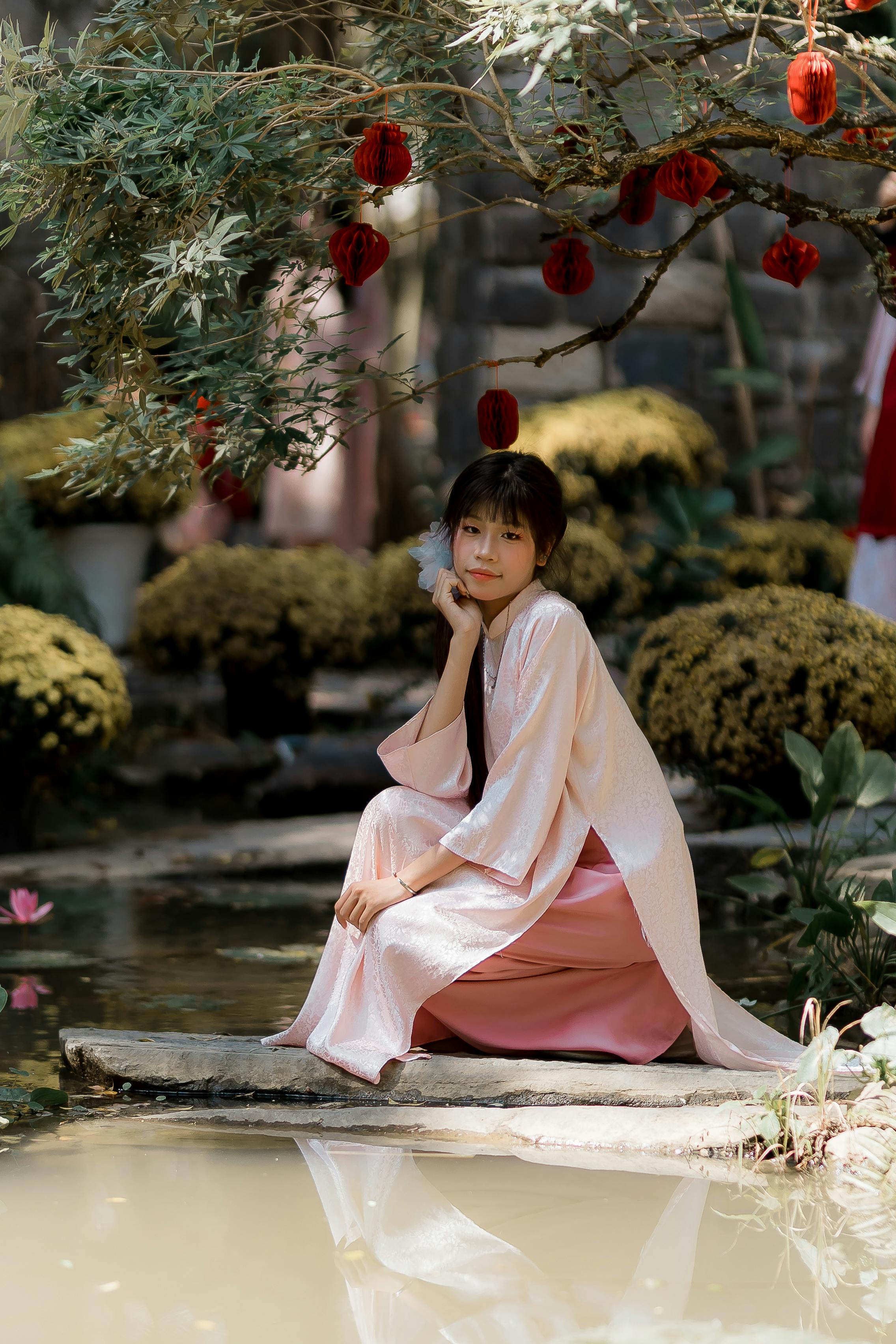 Ilmainen Perinteiseen asuun pukeutunut nuori nainen istuu kauniisti rauhallisen lammen rannalla, jonka yläpuolella loimuavat punaiset lyhdyt. Kuvapankkikuva