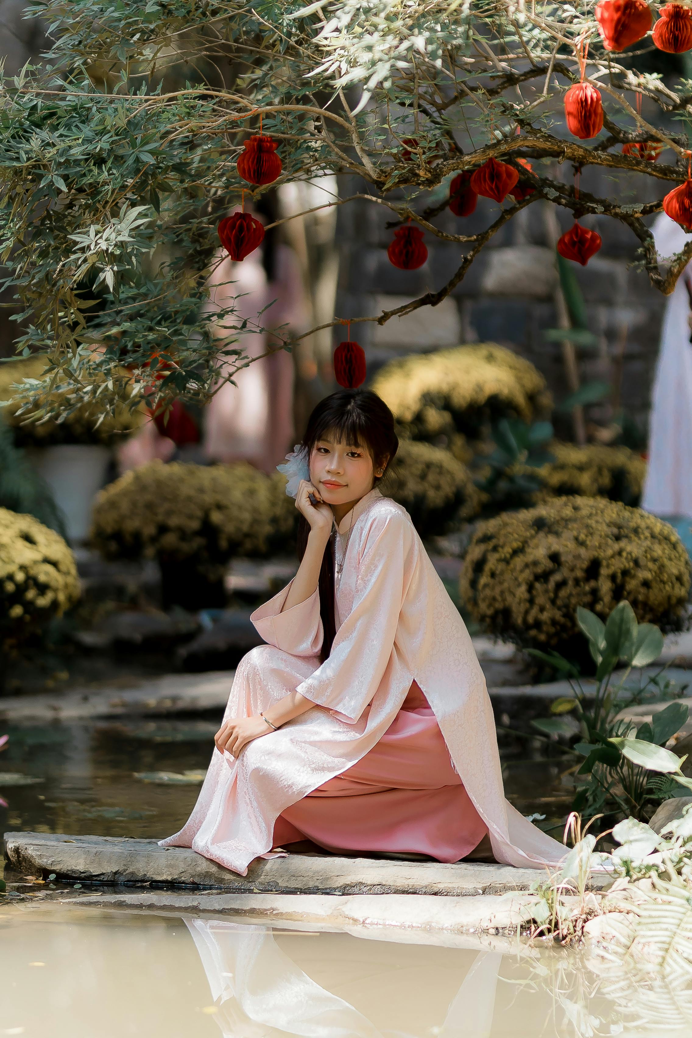 A young woman in traditional attire poses gracefully by a serene pond.