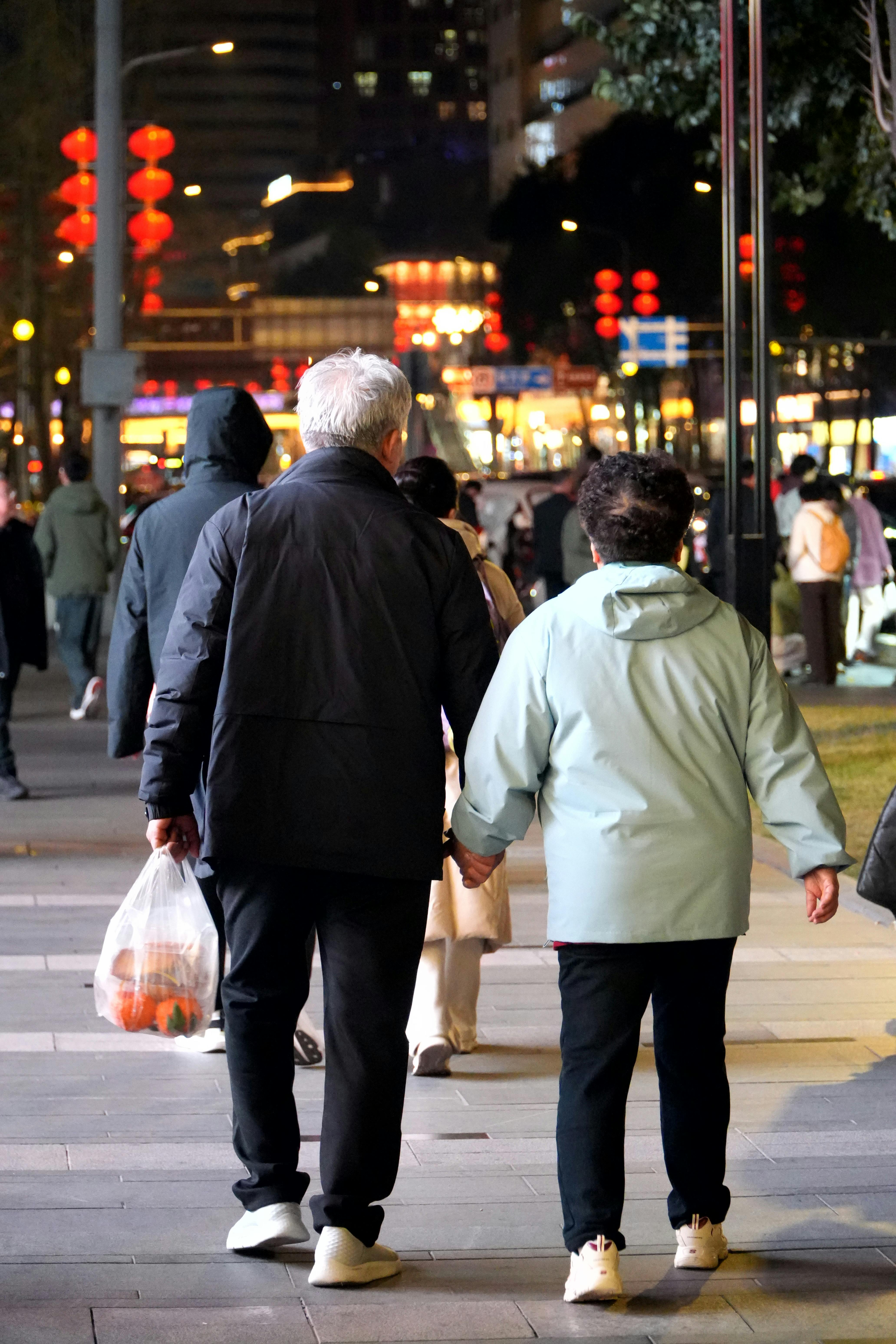 ฟรี คู่รักสูงอายุจับมือกันเดินลงไปตามถนนที่พลุกพล่านในเวลากลางคืนท่ามกลางแสงไฟของเมือง คลังภาพถ่าย
