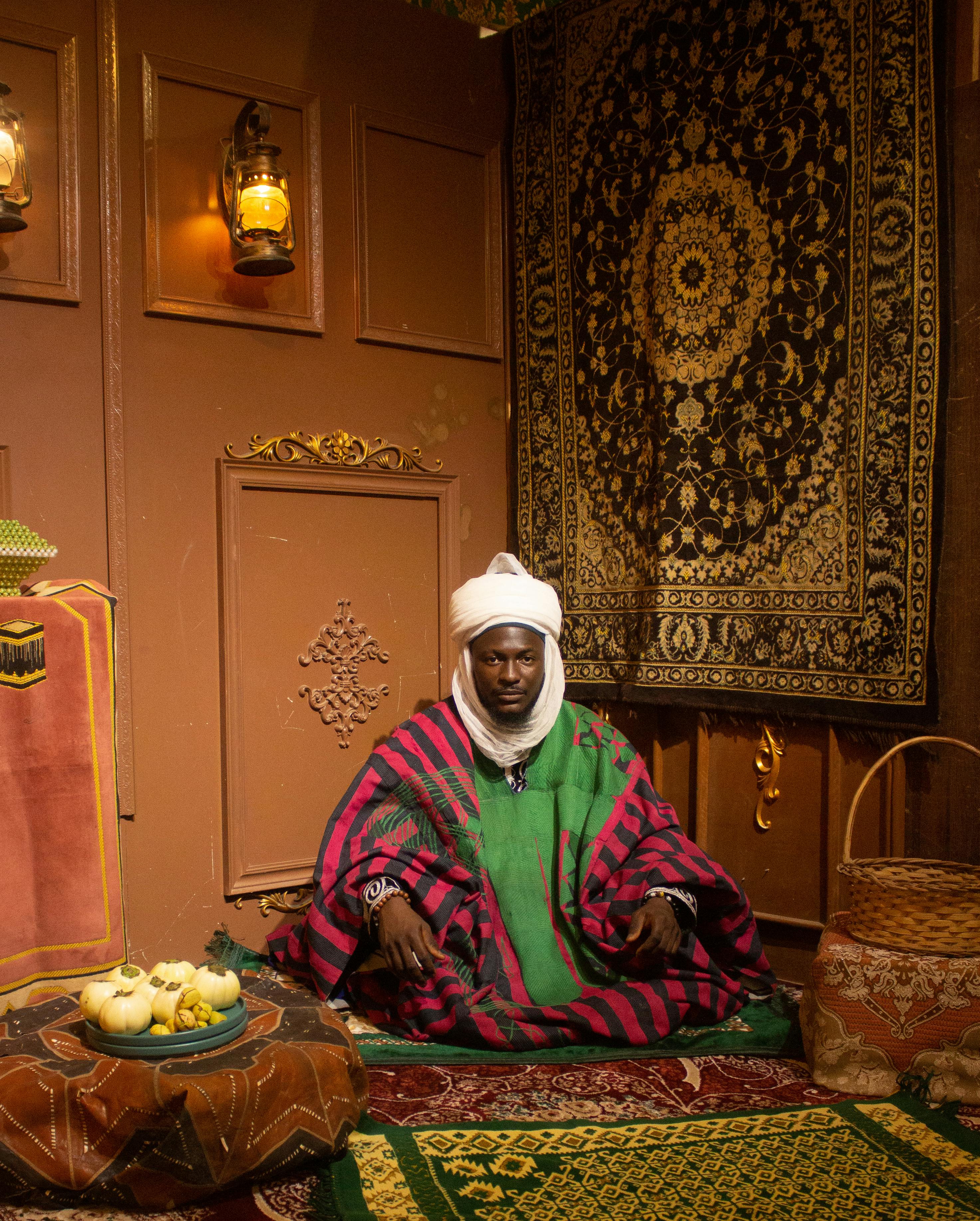 ฟรี ชายในชุดพื้นเมืองนั่งอยู่ในห้องที่ตกแต่งอย่างหรูหราด้วยพรมแขวนผนังที่วิจิตรงดงาม คลังภาพถ่าย