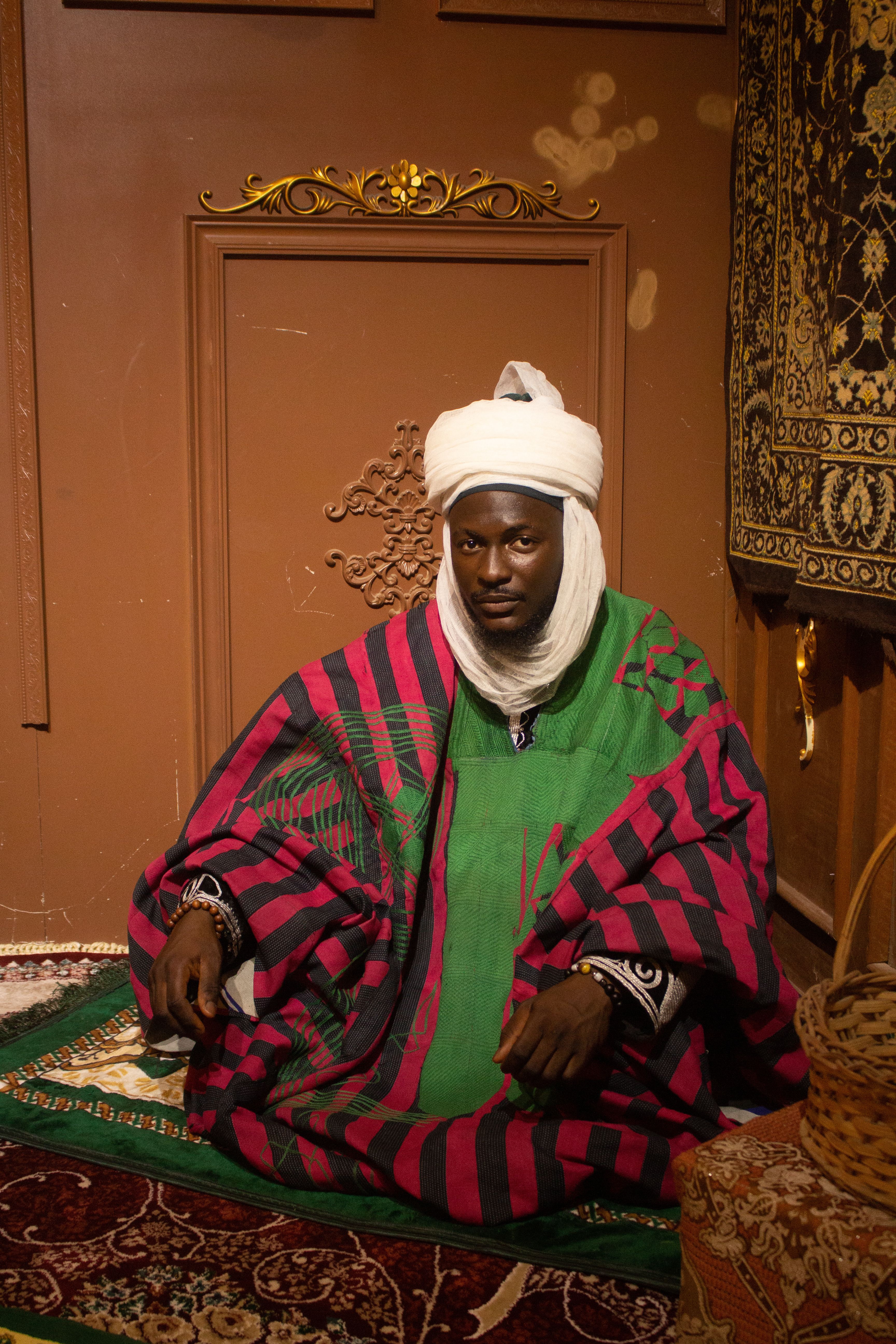 A man wearing traditional attire sits in an ornate room decorated with tapestries.