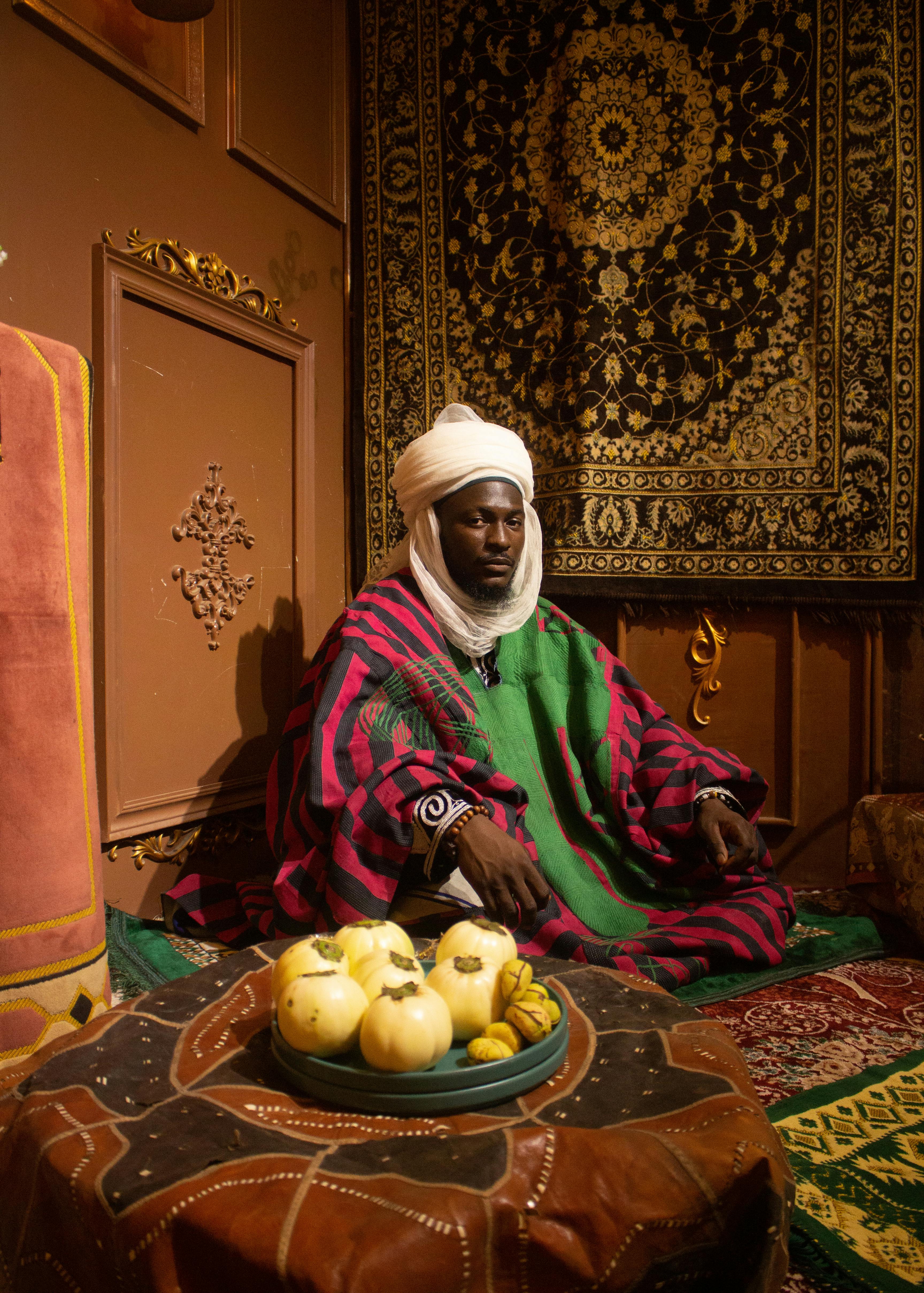 ฟรี ชายในชุดพื้นเมืองสีสันสดใส นั่งอยู่ในห้องที่ตกแต่งอย่างหรูหราด้วยของประดับตกแต่งแปลกตาและผลไม้ คลังภาพถ่าย