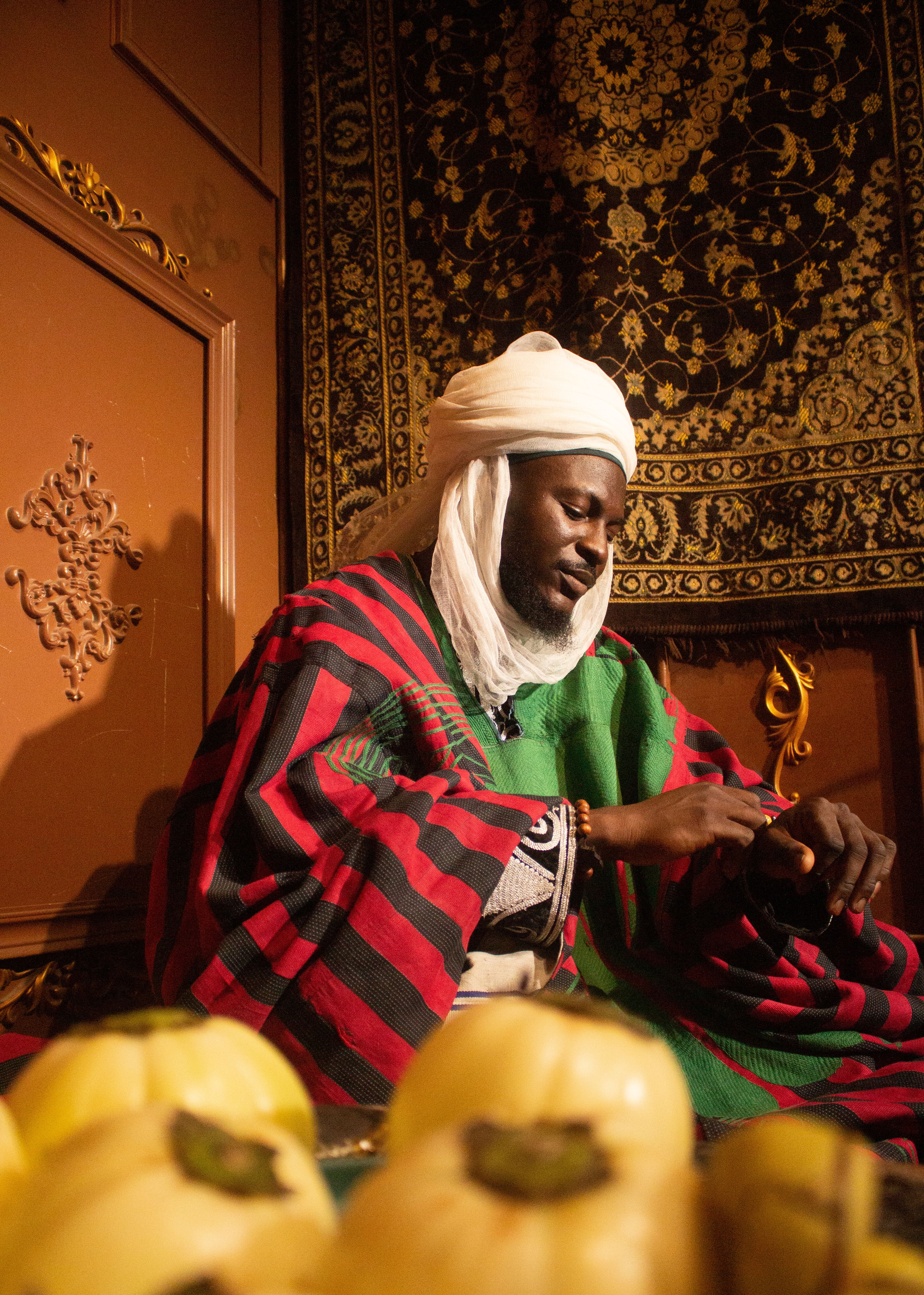 Portrait of African man wearing traditional clothing indoors, with decorative background and fruit.