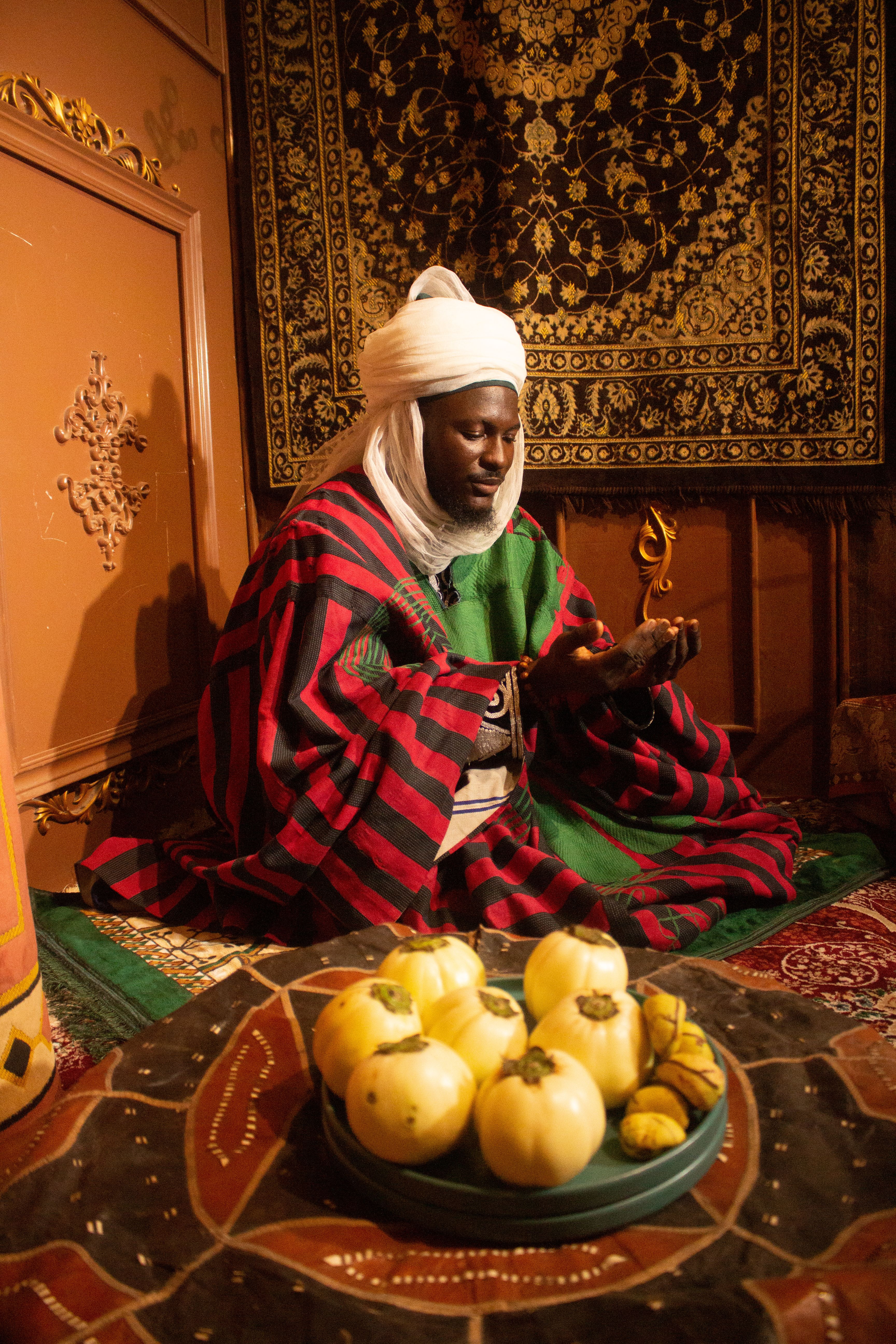 An African man in traditional attire sits in a richly decorated room with cultural items around him.