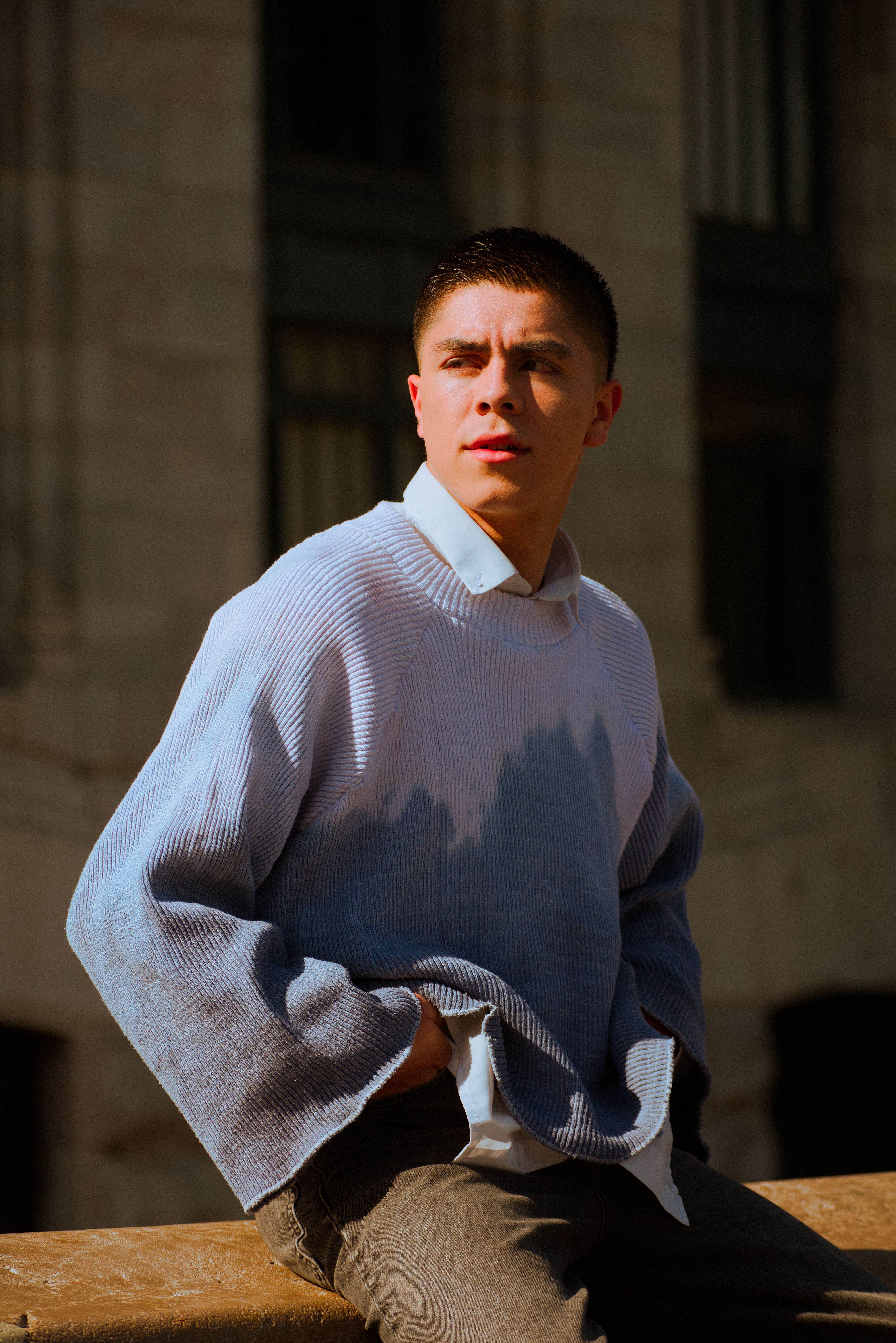 Young man wearing a blue sweater posing outdoors in daylight.