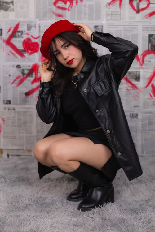Free Stylish woman in a red beret, black leather jacket, and black boots posing indoors with a creative newspaper backdrop. Stock Photo