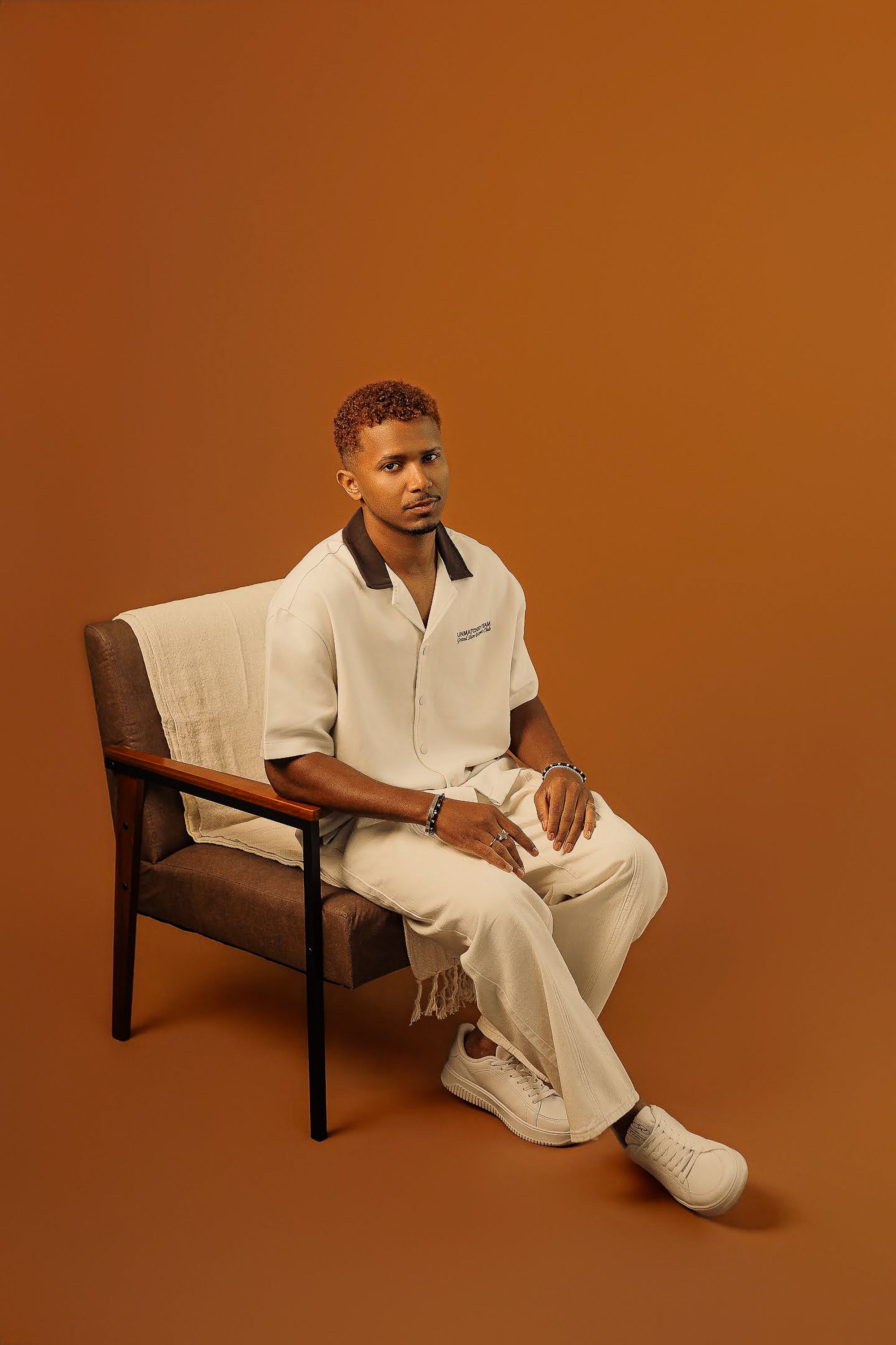 A fashionable man wearing white clothing poses in an orange studio setting.