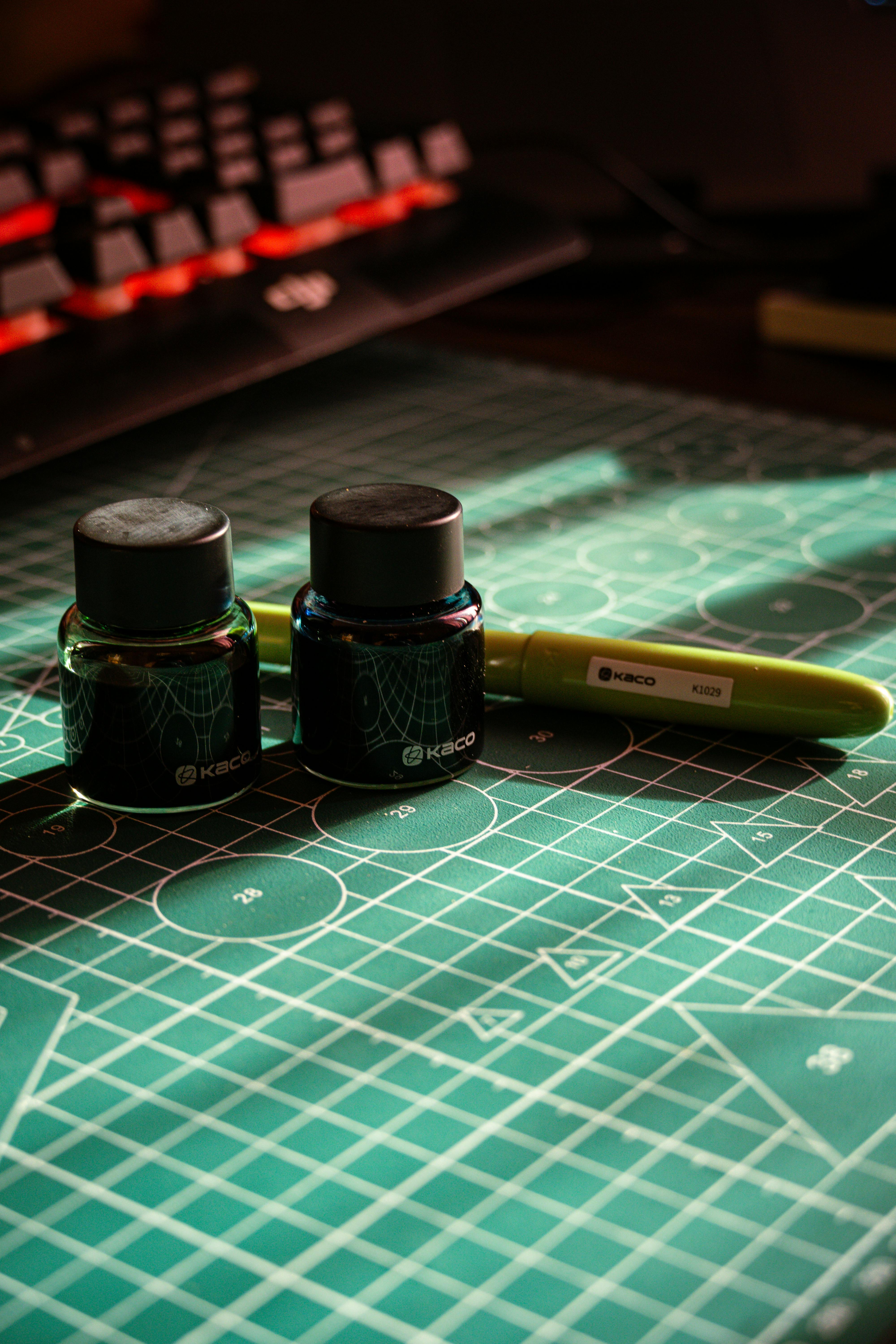 bezplatná Dvě lahvičky s inkoustem a zelené pero na stole s řezací podložkou pod teplým osvětlením. Základová fotografie