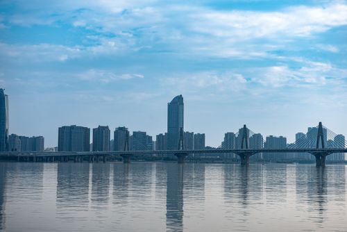 Free A panoramic view of a modern city skyline featuring a bridge over calm water under a blue sky. Stock Photo