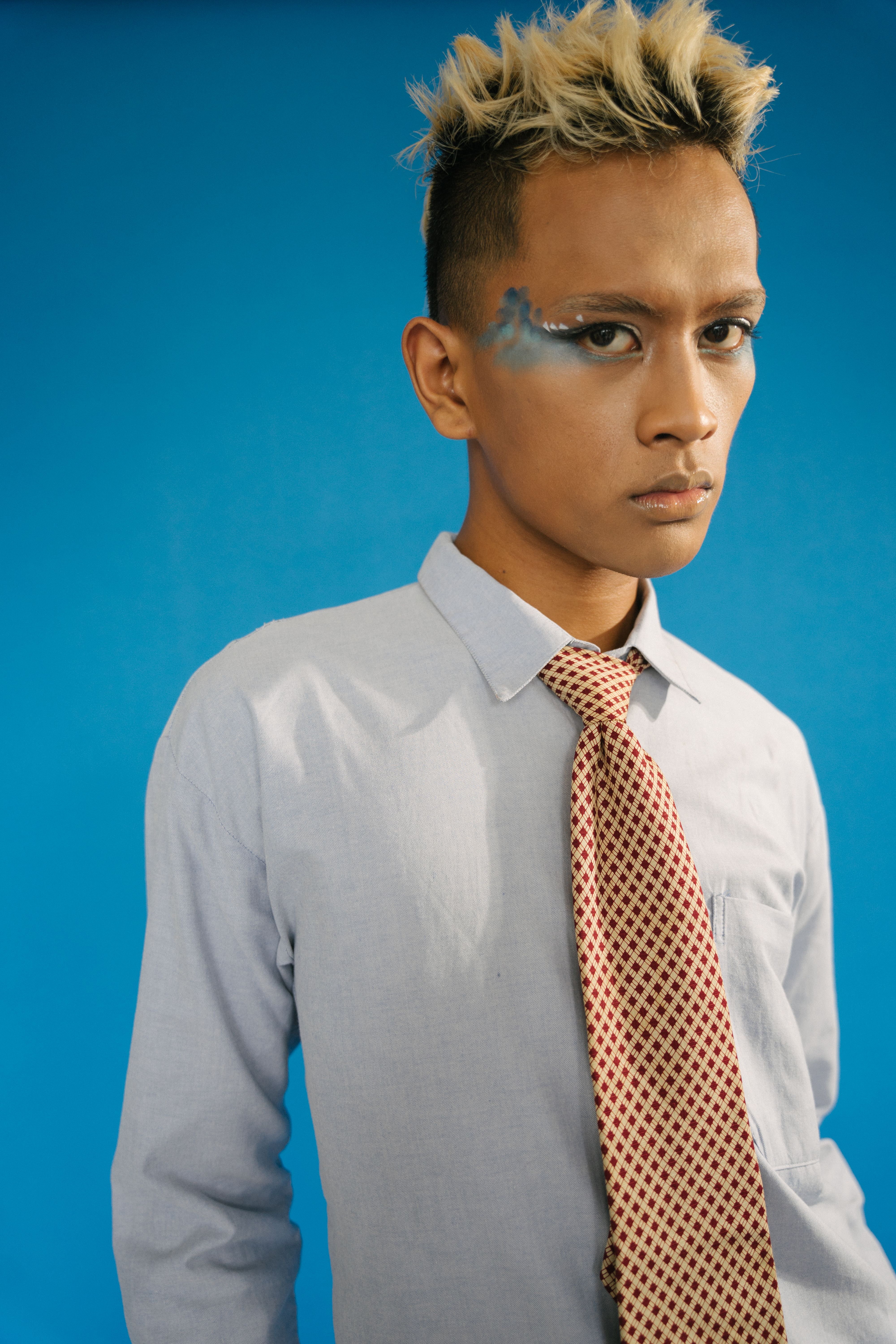 A young adult with artistic makeup, wearing a patterned tie against a vibrant blue backdrop.