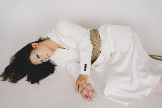 A conceptual portrait of a woman in white attire tied with rope, expressing vulnerability and emotion.