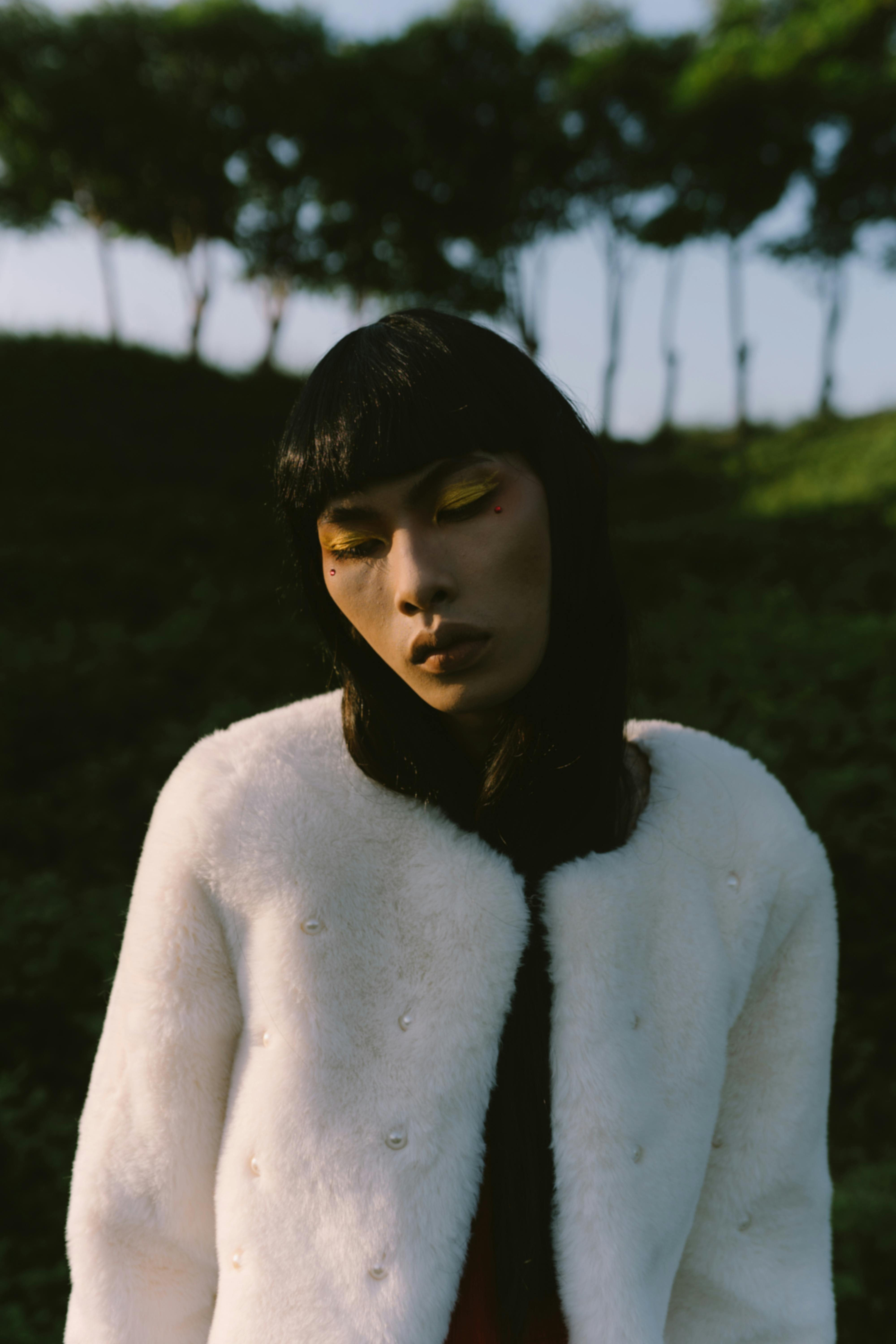 Miễn phí Một người phụ nữ mặc áo khoác trắng với lớp trang điểm mắt màu vàng nổi bật đứng ngoài trời, xung quanh là cây cối. Ảnh lưu trữ