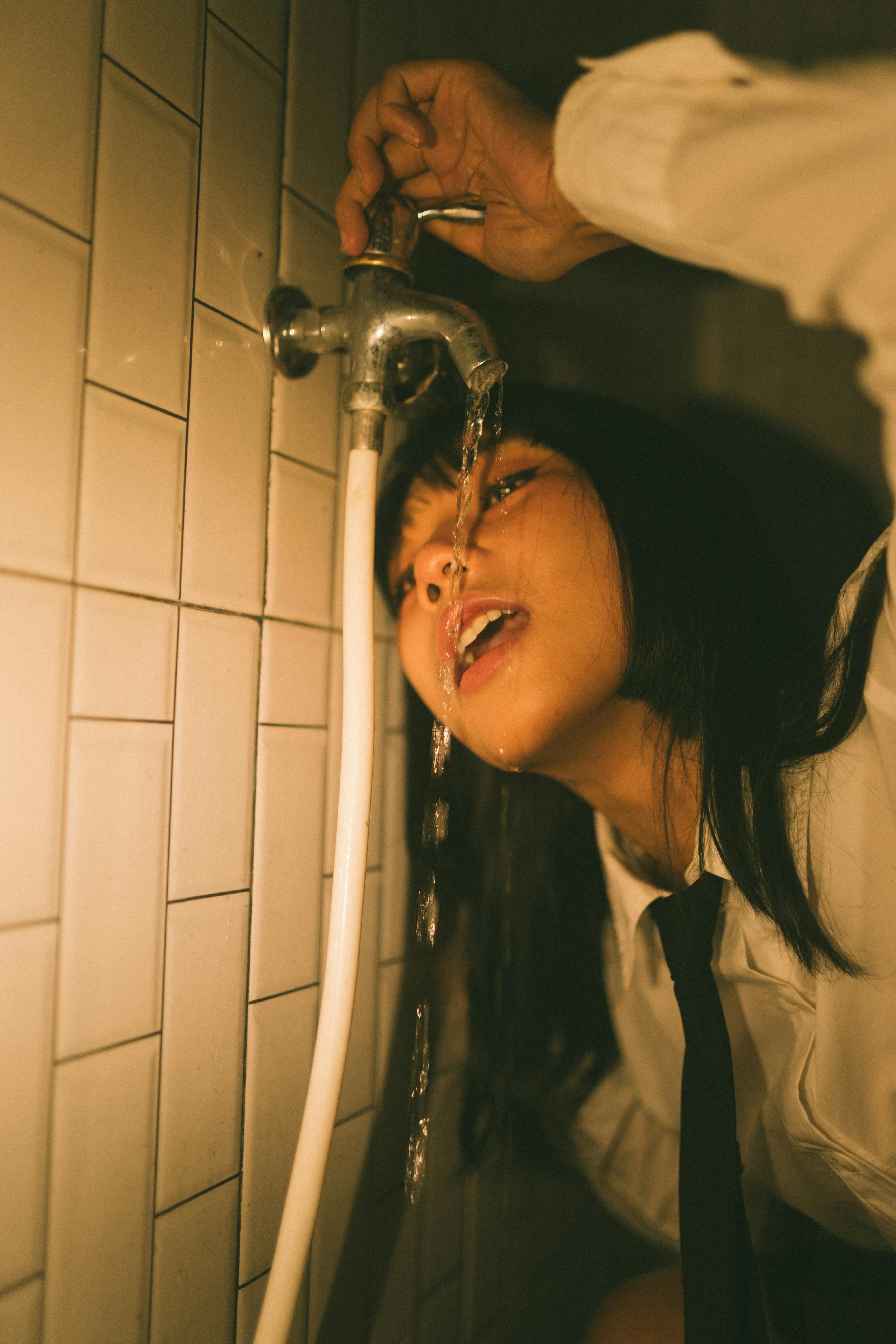 Miễn phí Một cô gái trẻ uống nước từ vòi trong một căn phòng lát gạch thiếu ánh sáng, tạo nên một bầu không khí độc đáo và mang tính nghệ thuật. Ảnh lưu trữ