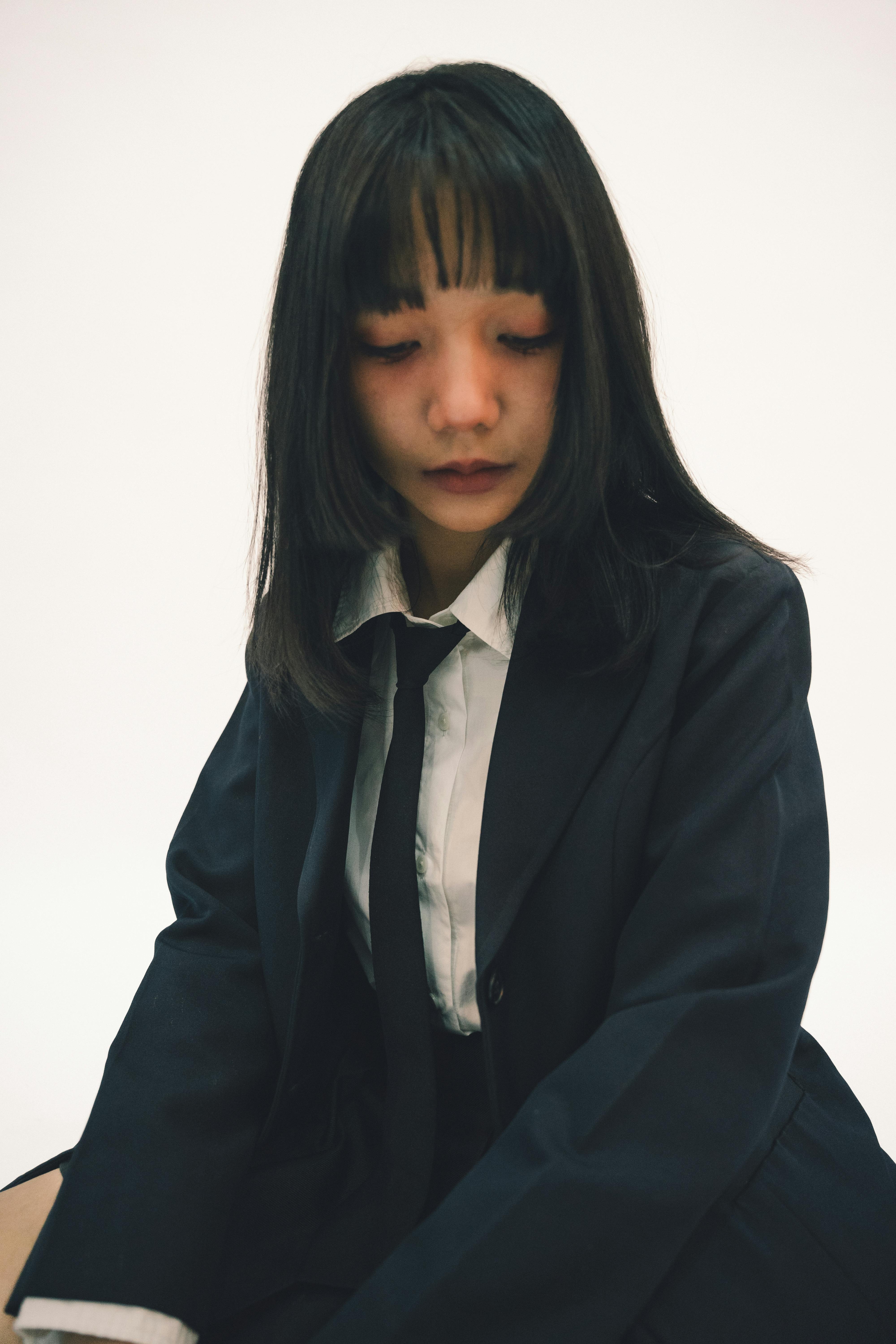 A young woman in a school uniform gazes down with a thoughtful expression, set against a white background.