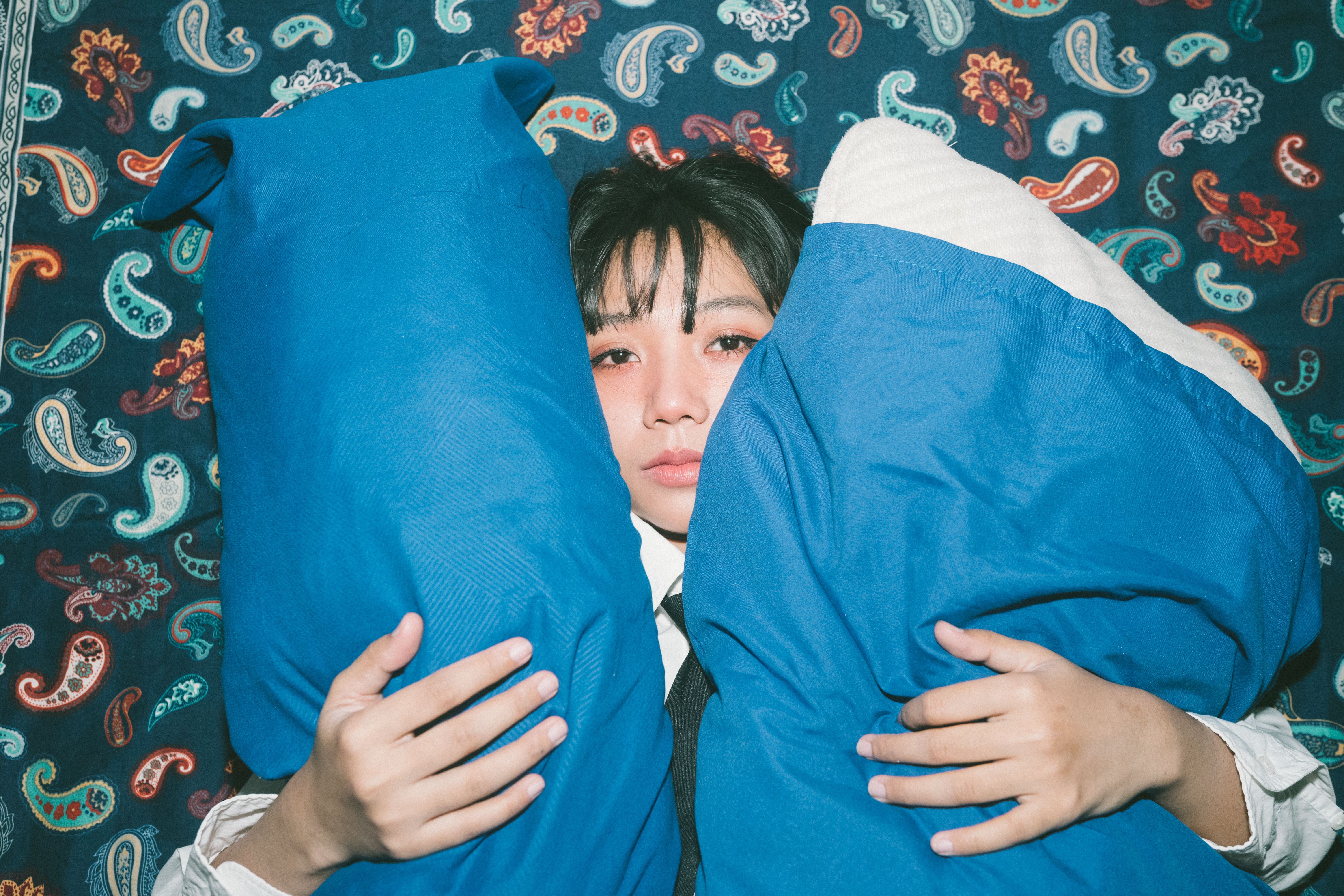 A young adult holding blue pillows against a vibrant paisley backdrop indoors.