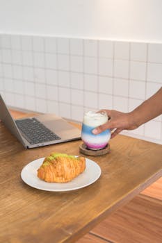A colorful latte and croissant on a wooden desk with a laptop. Ideal for modern workspace themes.