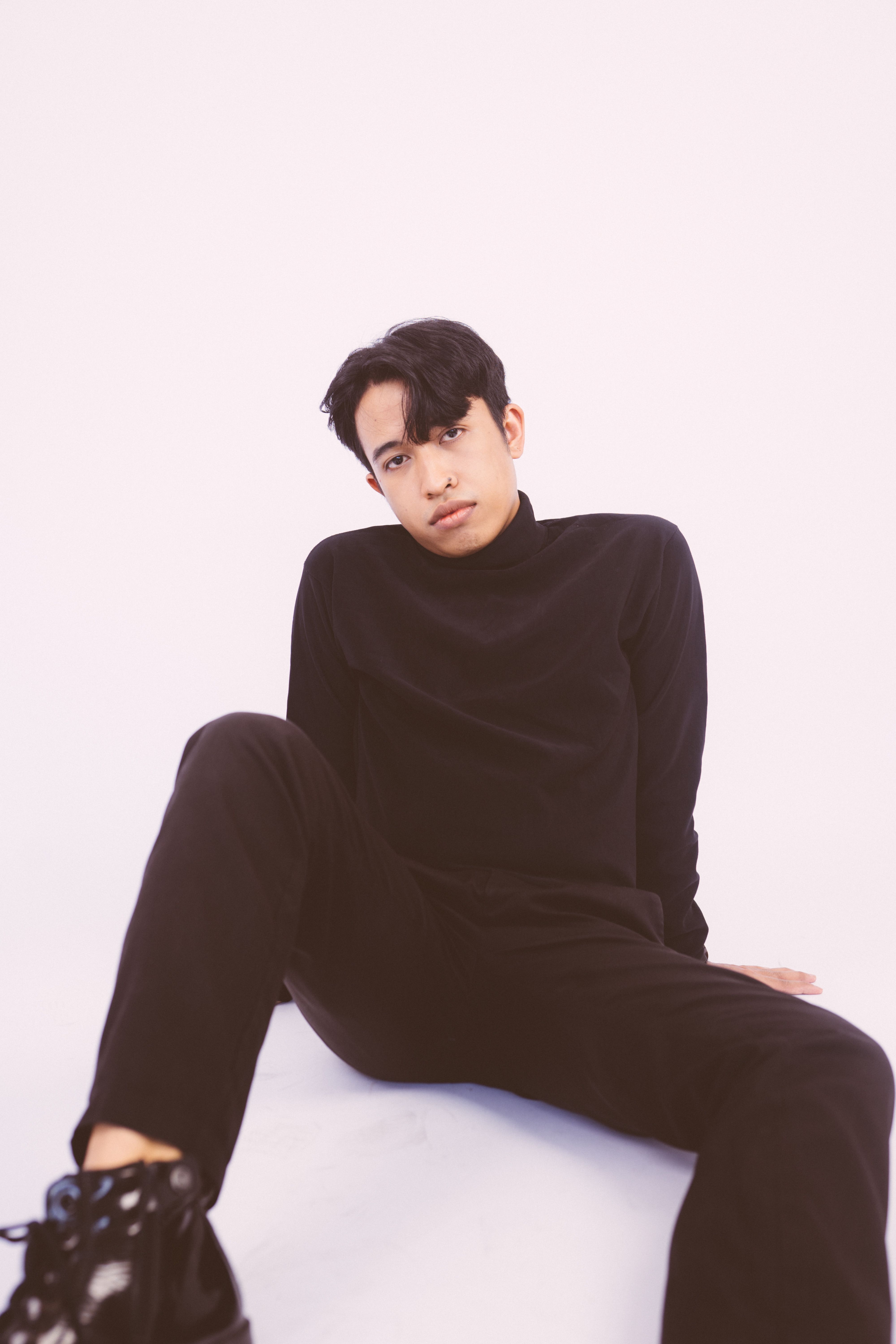 Young man in black attire poses casually against a minimalist white backdrop.