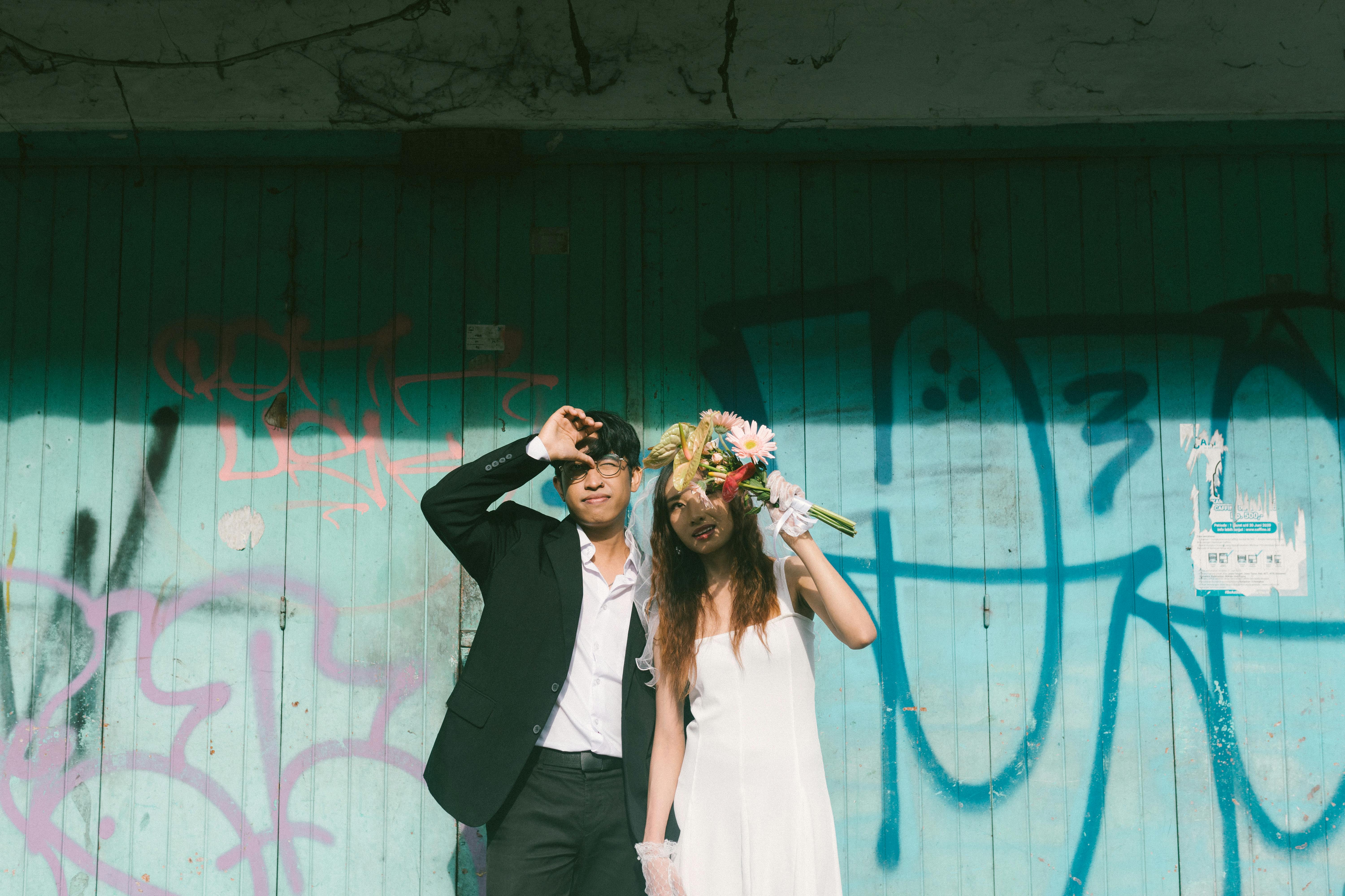 Ücretsiz Şık bir çift, rengarenk grafiti duvarının önünde buketleriyle poz veriyor. Modern şehir havası. Stok Fotoğraflar