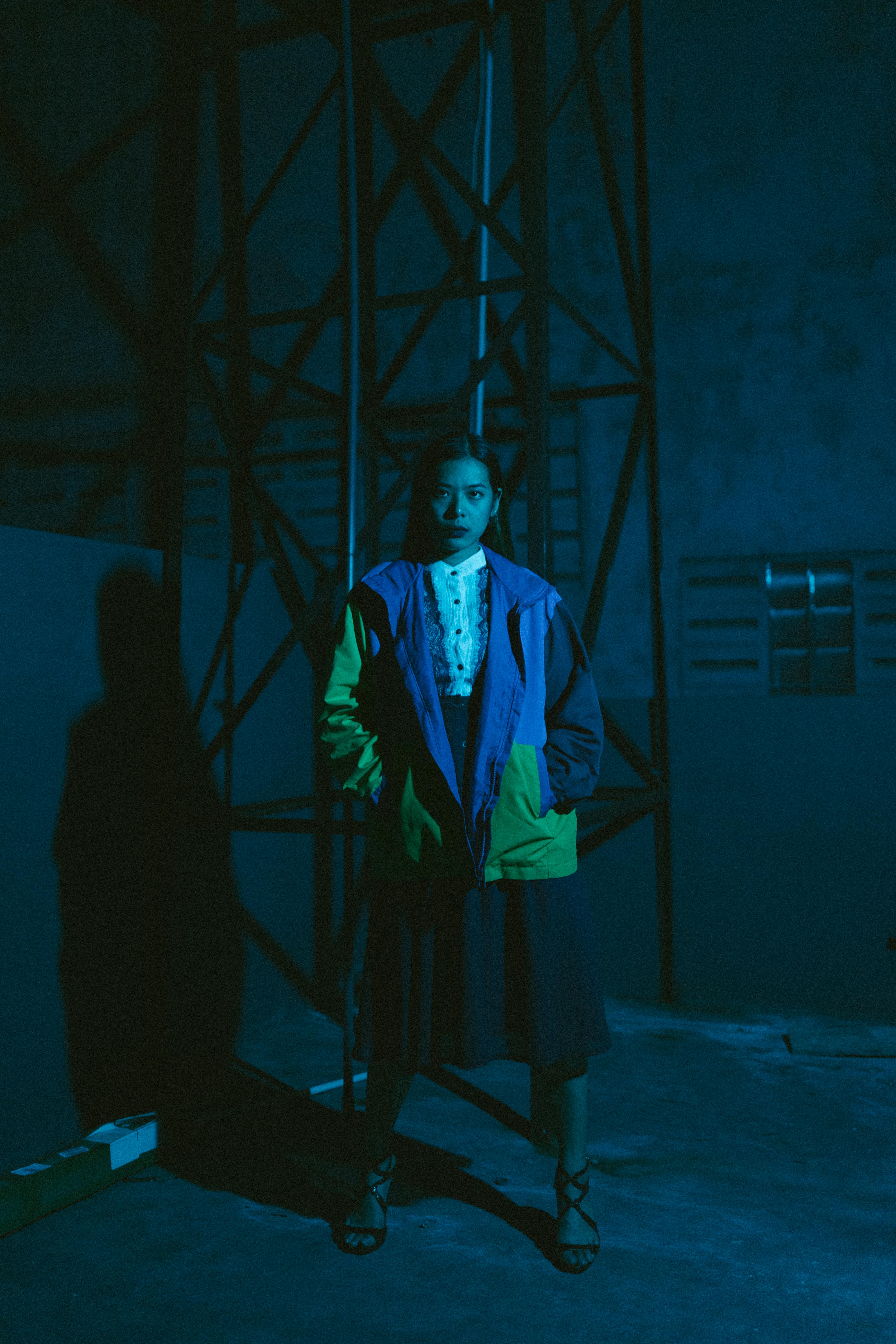 Free A young woman stands in an industrial environment with dramatic lighting and shadows emphasizing her colorful jacket. Stock Photo