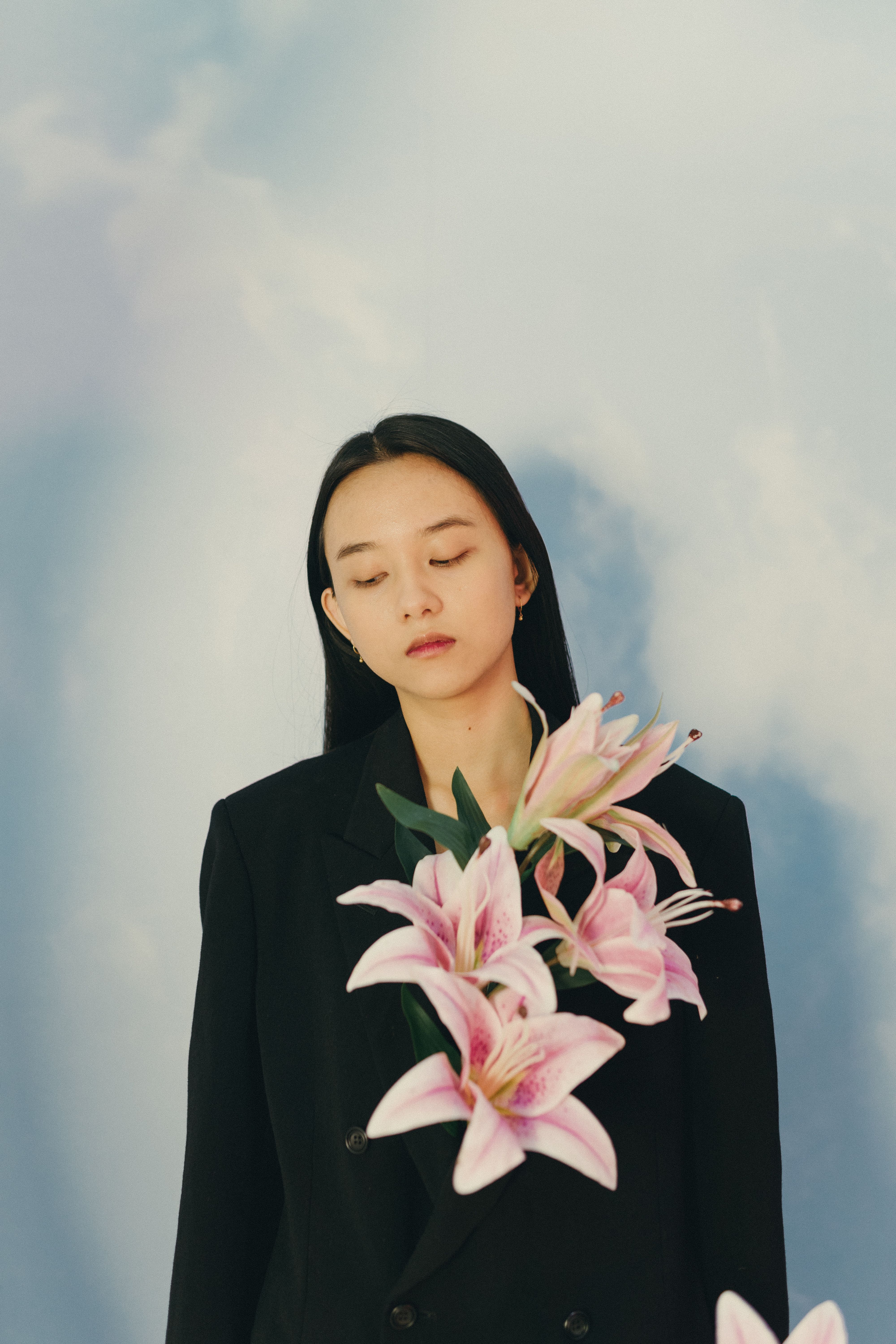 Serene portrait of a woman in a black blazer adorned with pink lilies against a soft blue background.
