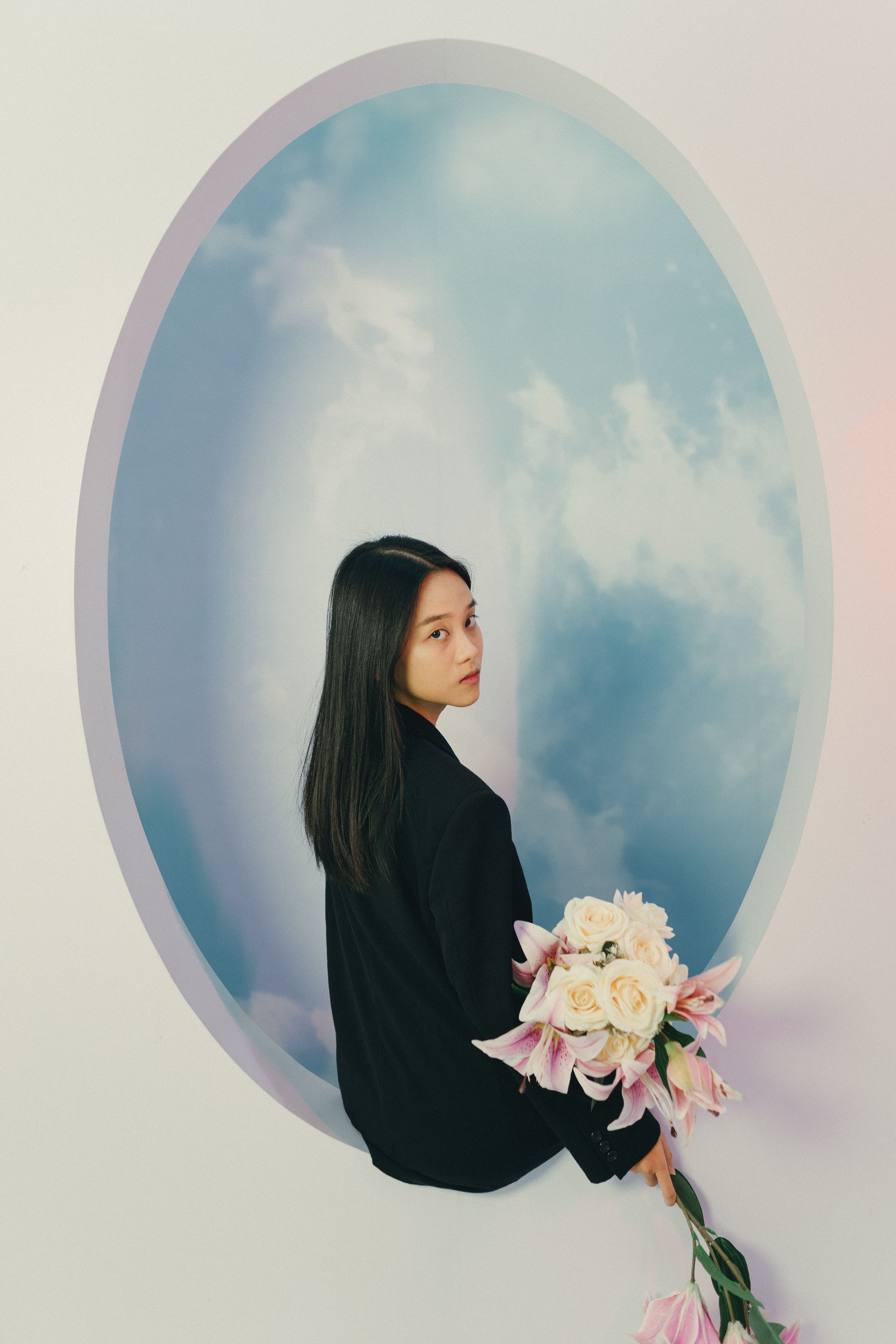 A woman holding flowers stands against a sky-themed backdrop, creating a dreamy artistic portrait.