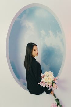 A woman holding flowers stands against a sky-themed backdrop, creating a dreamy artistic portrait.