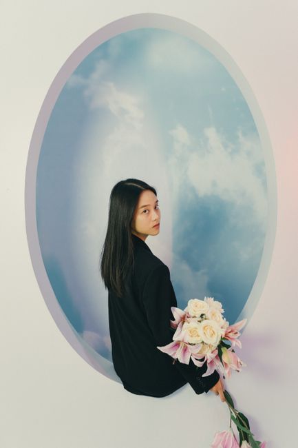 A woman holding flowers stands against a sky-themed backdrop, creating a dreamy artistic portrait.