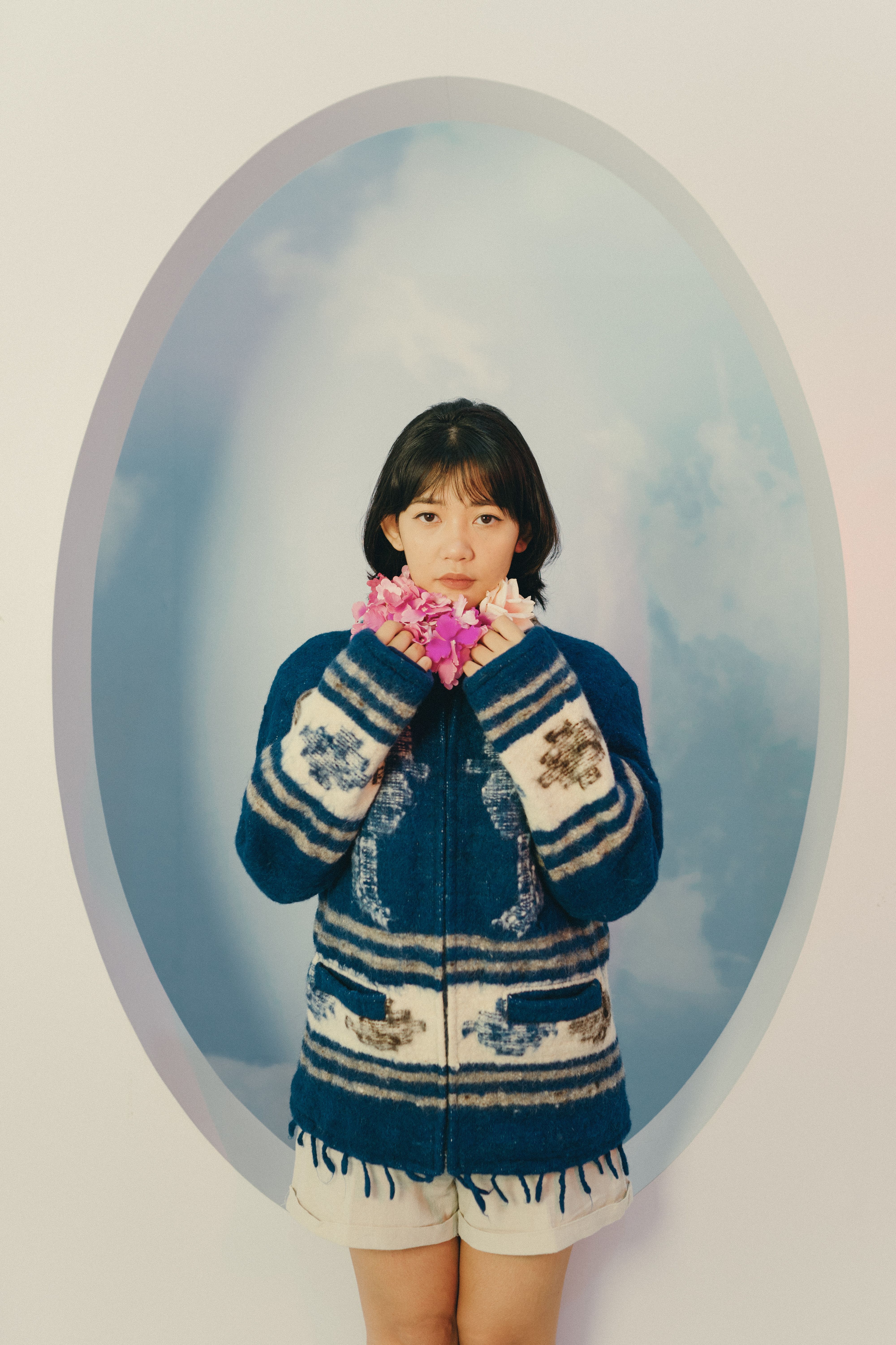 Woman in a blue and white sweater holds vibrant flowers against a pastel backdrop.