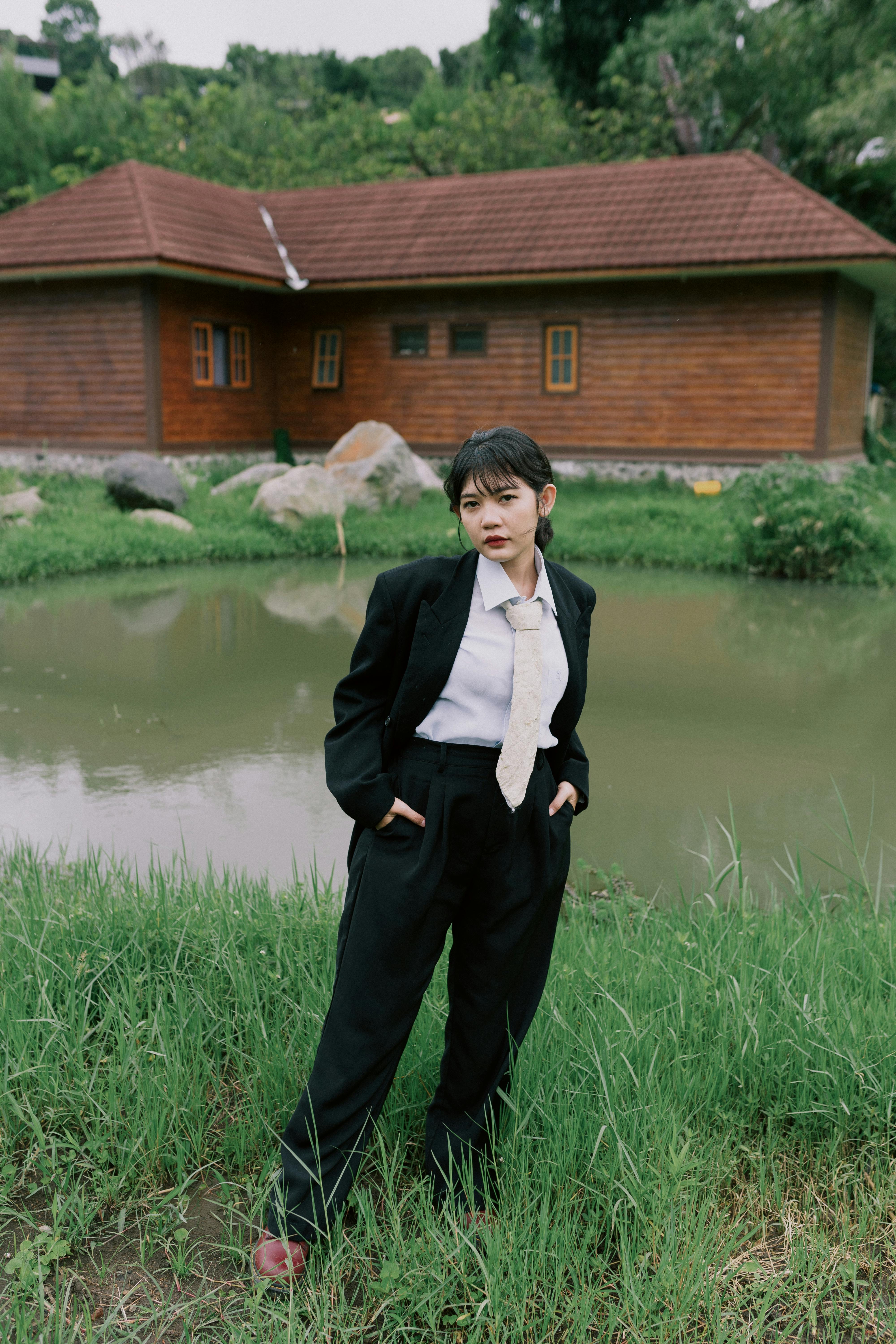 Ilmainen Itsevarma nuori nainen muodollisessa mustassa puvussa seisoo lammen rannalla puutalon edessä. Kuvapankkikuva