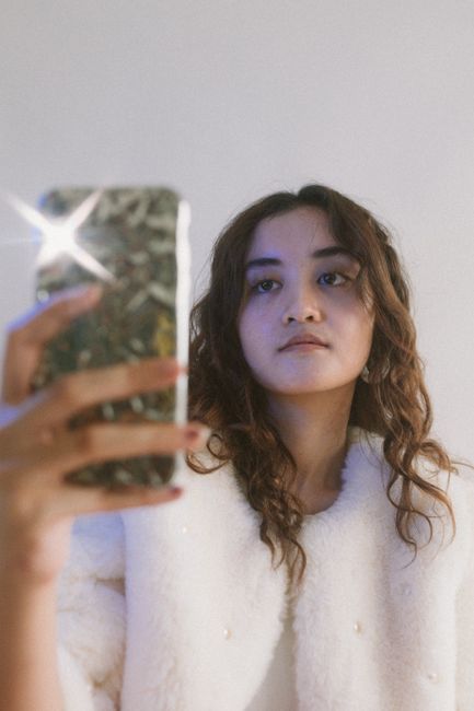 Young woman capturing a selfie with a stylish phone, wearing a plush white coat against a neutral background.