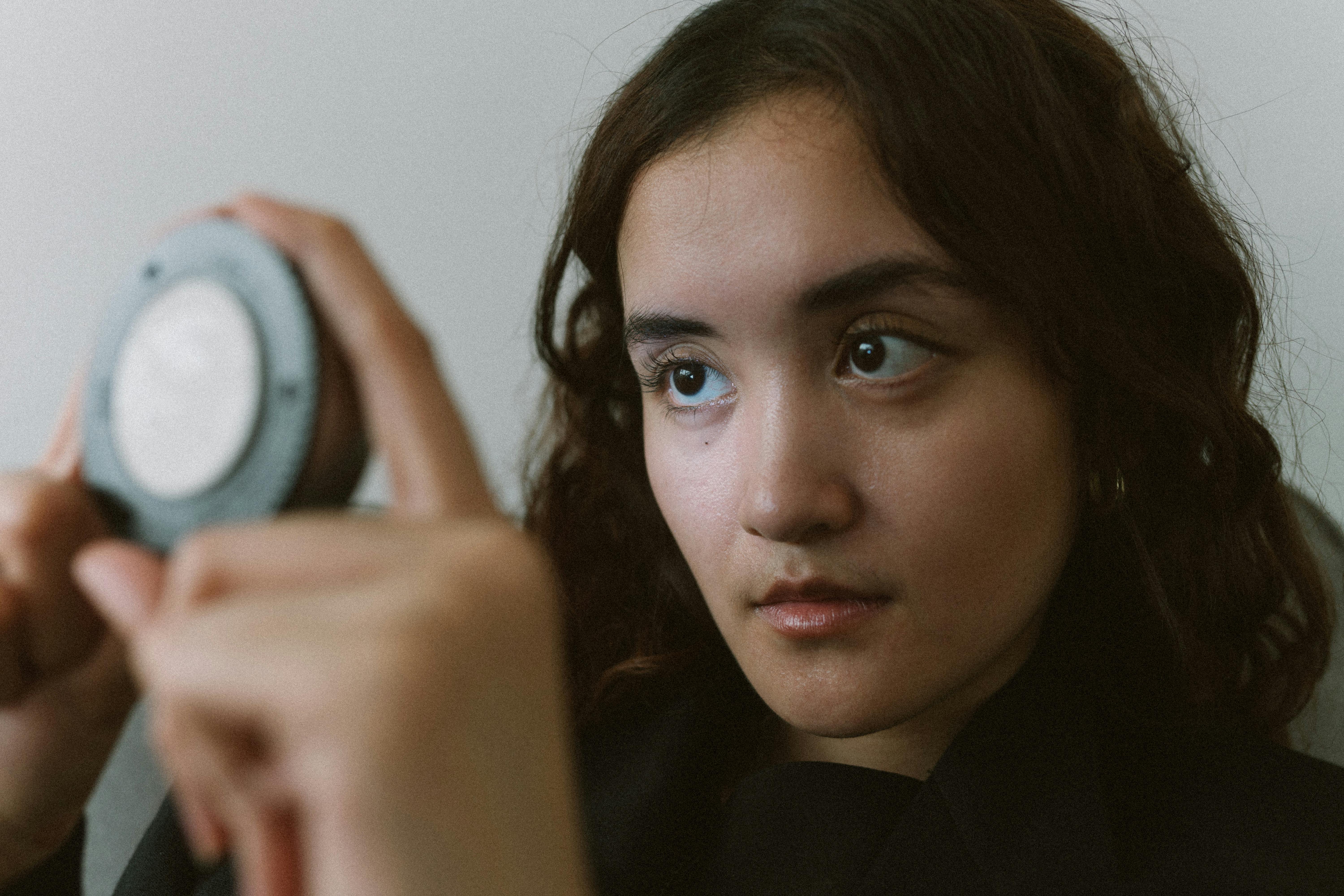 bezplatná Mladá žena pozorně hledící do kompaktního zrcadla uvnitř, zobrazující sebereflexi. Základová fotografie