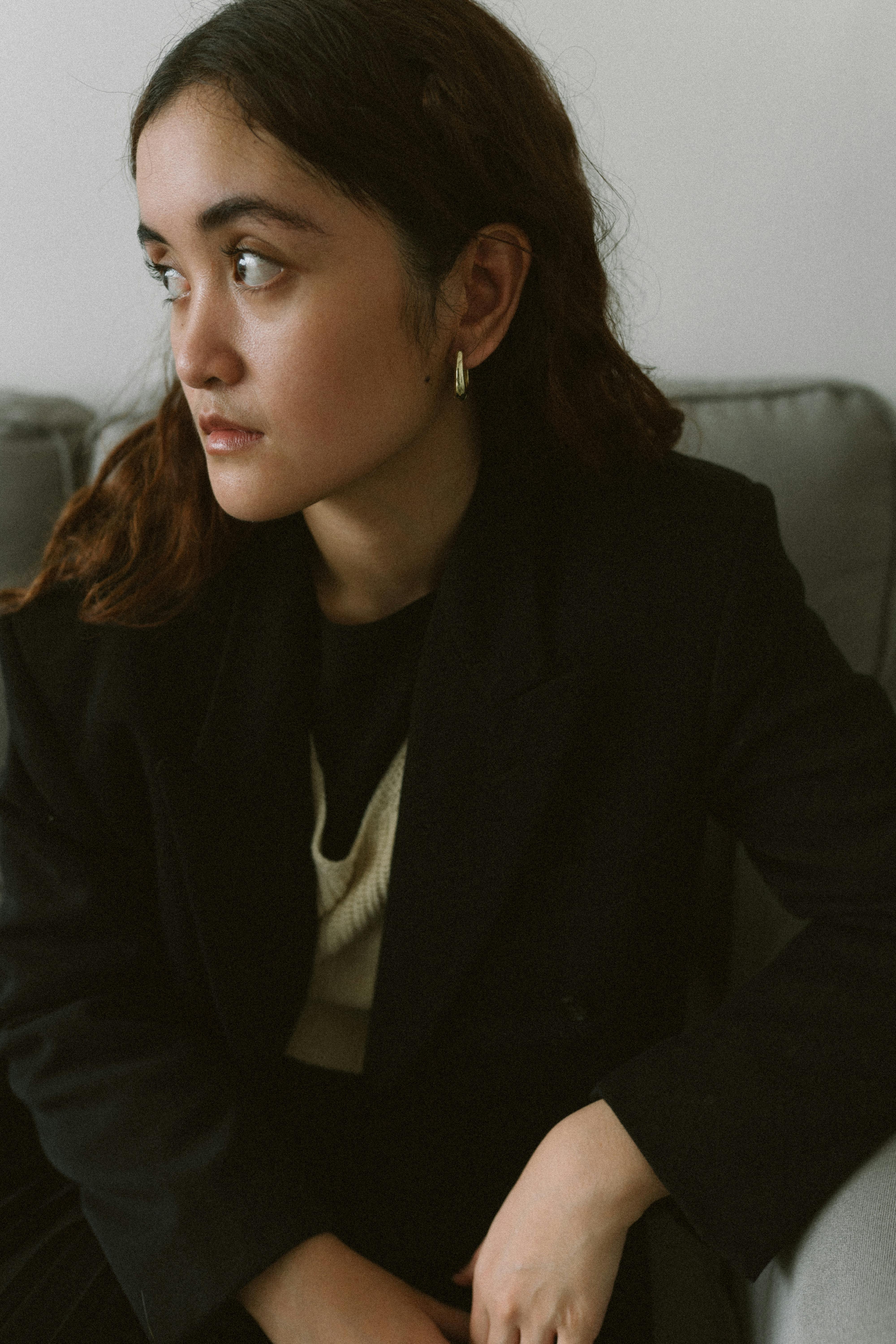 Bezpłatne Portret kobiety w czarnym żakiecie siedzącej w pomieszczeniu z zamyślonym wyrazem twarzy. Zdjęcie z galerii