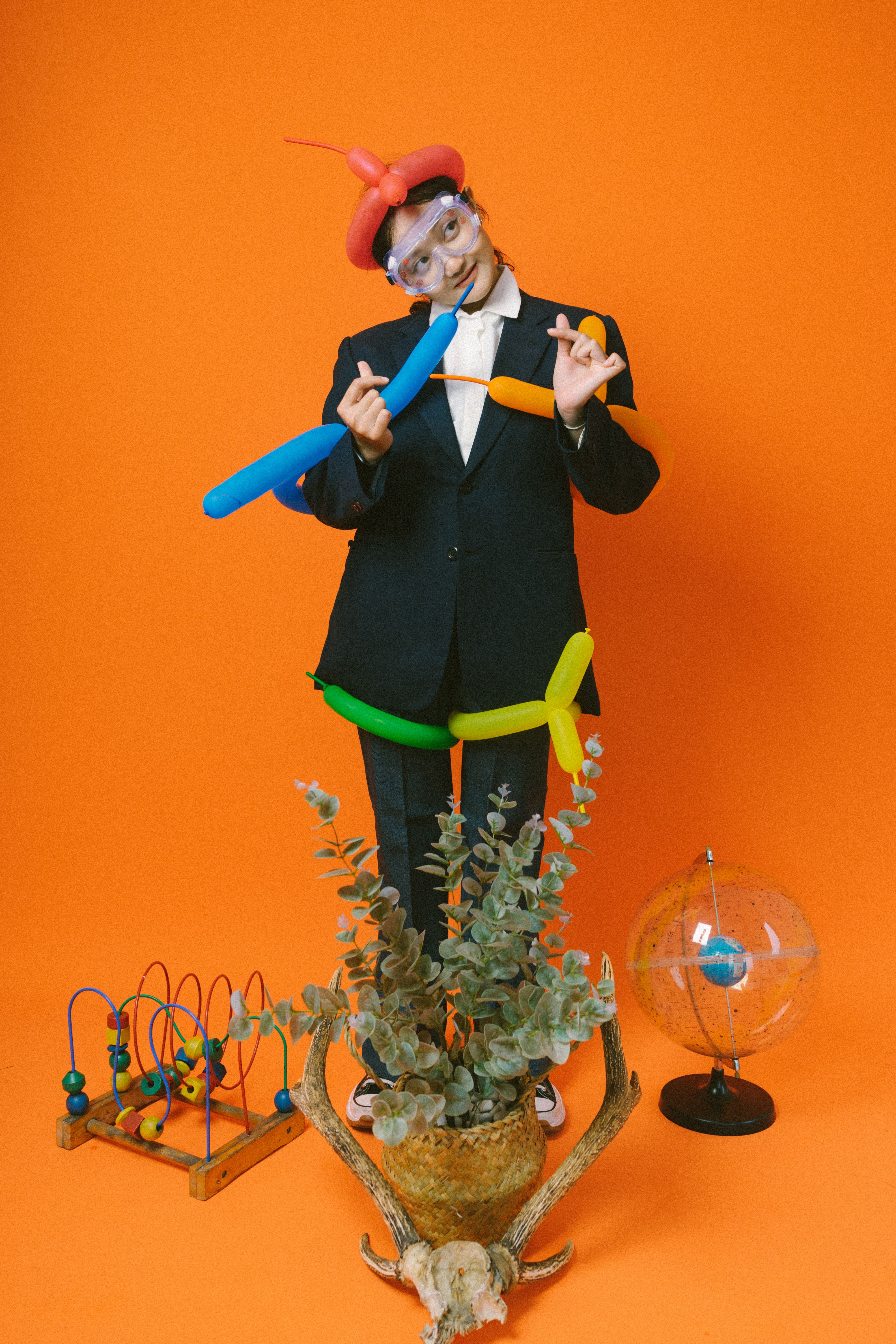 A playful individual in a suit with balloons, goggles, and a bright orange backdrop.