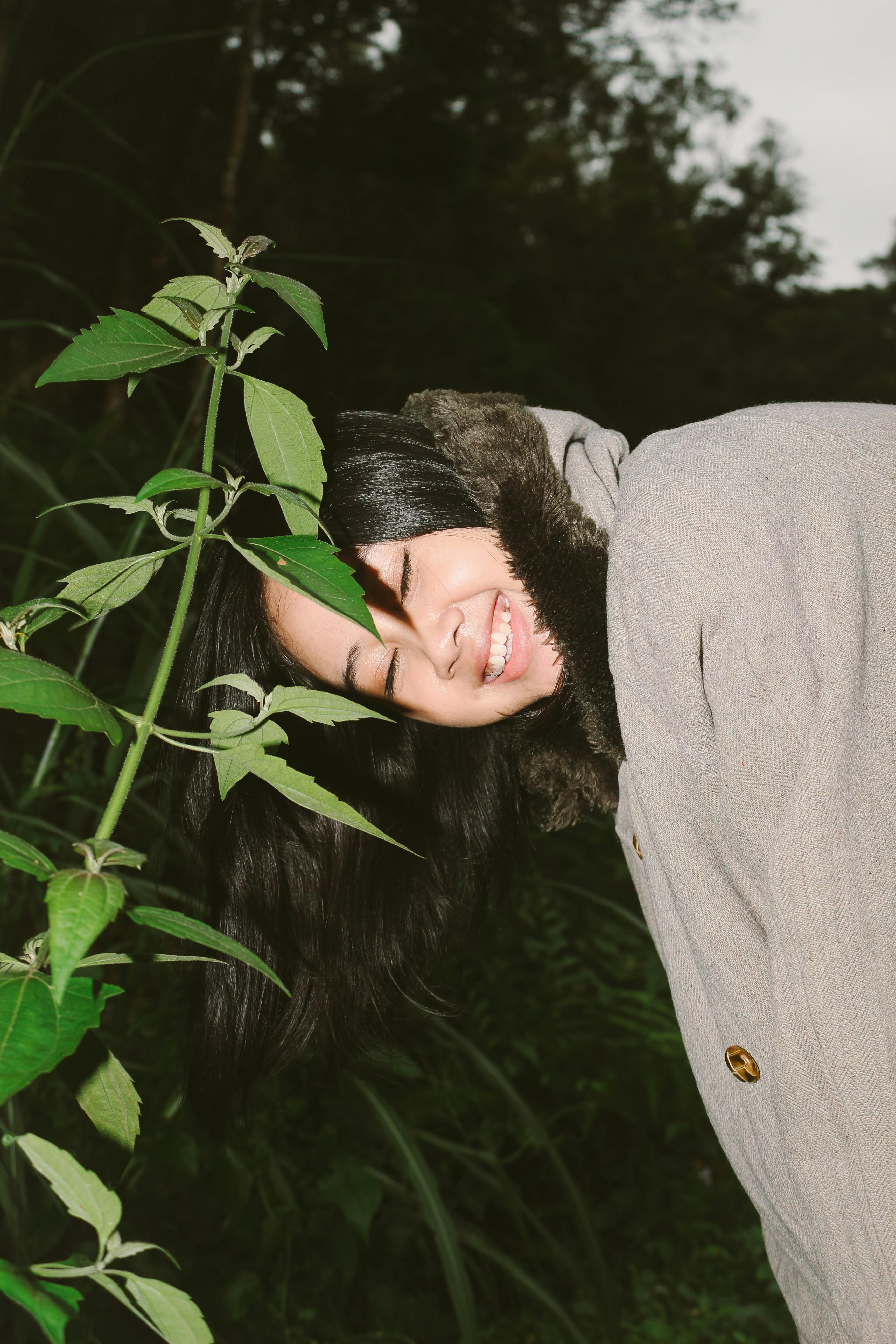 Ilmainen Takkiin pukeutunut nainen hymyilee ja nojaa leikkisästi lehtikasviin rehevän vihreässä ulkoympäristössä. Kuvapankkikuva